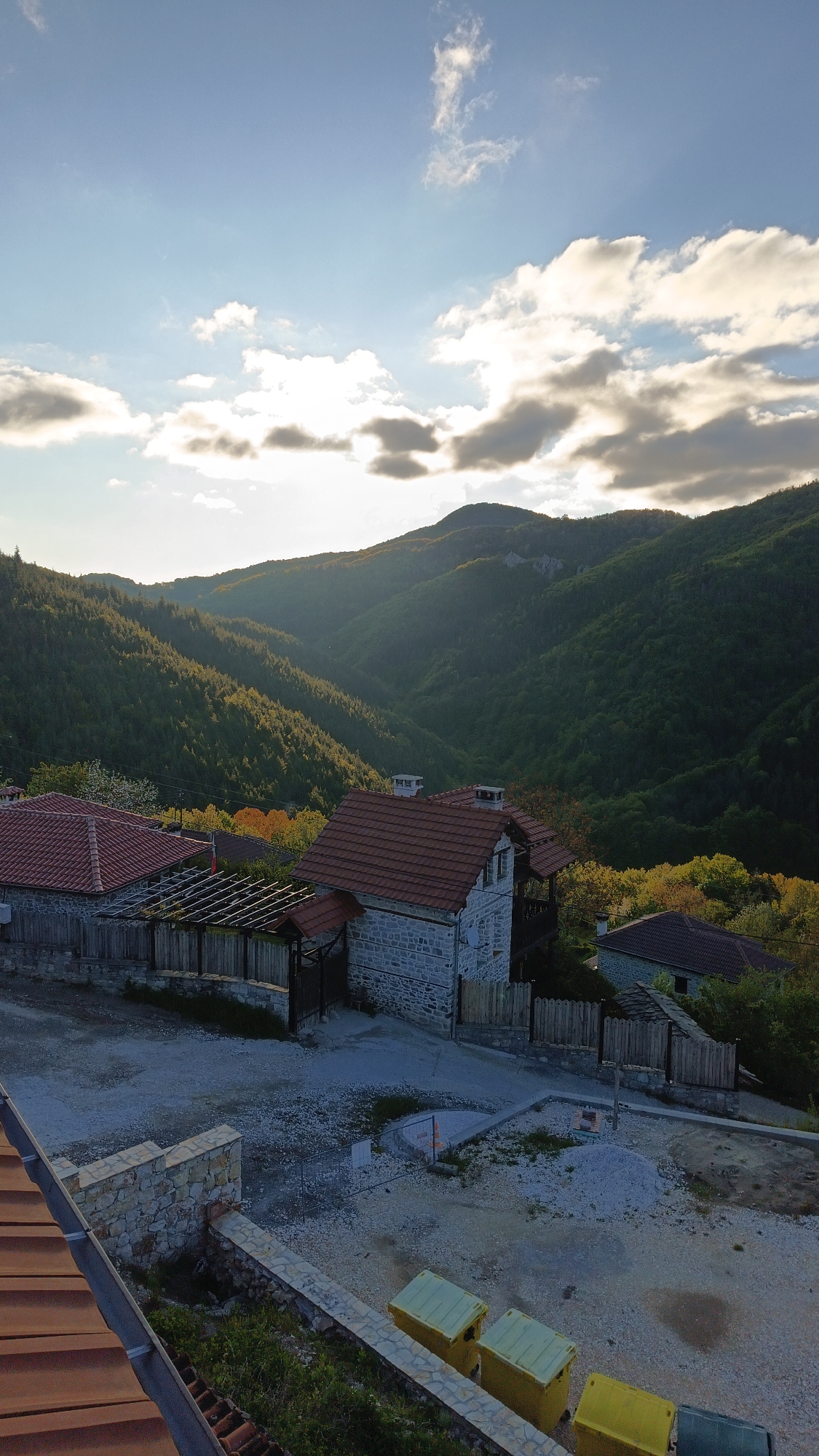 The image depicts a scenic view of what appears to be a village nestled in the mountains of Vrata, Bulgaria. The foreground features a construction site with piles of gravel and construction materials, as well as several yellow bins. A low stone wall separates the site from a lower-level roof with red tiles.
Beyond the construction area, there are stone houses with red tiled roofs, enclosed by wooden fences. The houses are set against a backdrop of lush, green mountains. The lighting suggests it is either early morning or late afternoon, with the sun partially obscured by clouds, creating a soft, diffused light across the landscape. The overall environment is rural and mountainous, suggesting a peaceful setting.