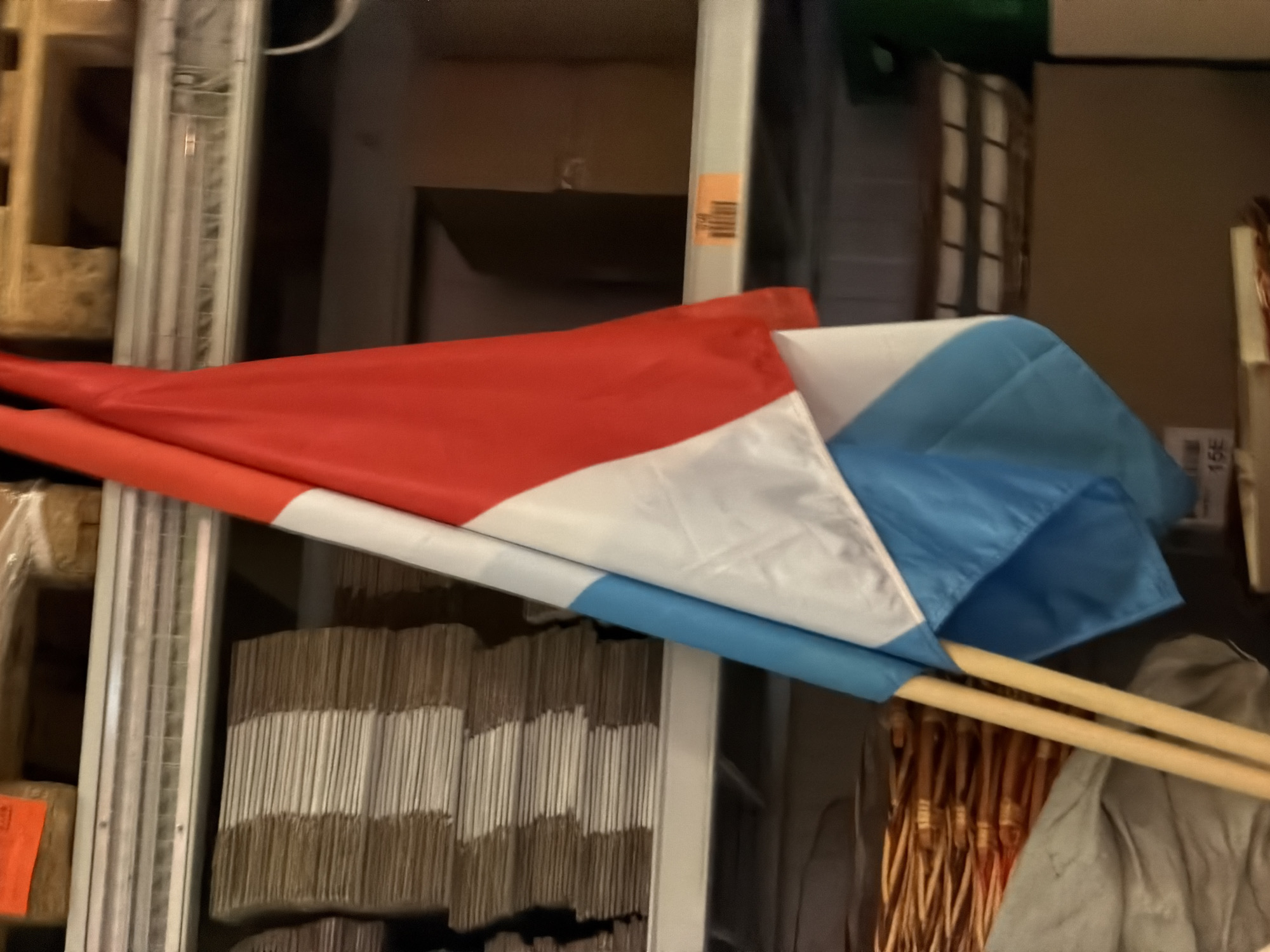 The image shows a close-up of a flag of Luxembourg, partially unfurled, with wooden poles extending downwards. The flag has three horizontal stripes: red, white, and light blue. In the background, there are shelves filled with stacked items, including what appear to be cardboard boxes and baskets. The scene suggests a storage or warehouse environment, possibly in Leudelange, Luxembourg, given the flag. The lighting is somewhat dim, with visible shelf structures and some orange labeling or stickers on the shelves.