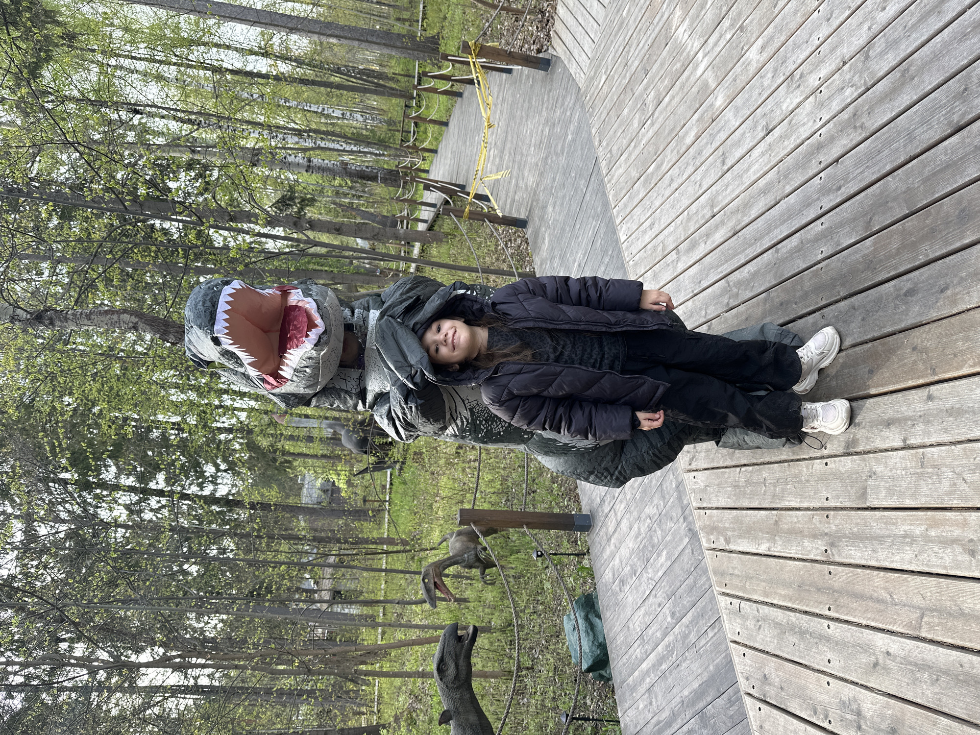 A young girl wearing a dark jacket, dark pants, and white sneakers is lying on a dinosaur statue with its mouth open on a wooden boardwalk. The scene is set at an outdoor park or attraction, featuring additional dinosaur statues and a backdrop of trees. There is a yellow safety rope in the background. The location is Izhevsk, Russia.