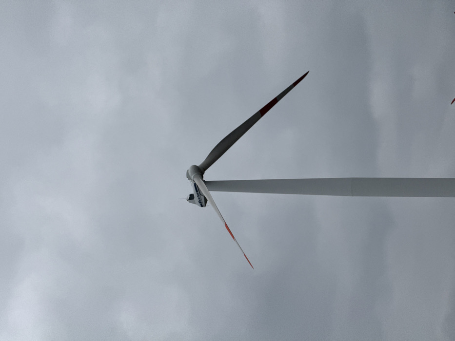 This image depicts a Vestas wind turbine located in Dornstadt, Germany, positioned against an overcast, uniform gray sky. The turbine consists of a white, tapering cylindrical tower supporting a white nacelle. The "Vestas" logo in blue text is visible on the side of the nacelle. Two rotor blades are visible; one extends upward and to the left from the nacelle, featuring a red tip, while the other extends downward and to the right, also with a red tip. The blades appear stationary. No people, other structures, or ground features are present in the frame.