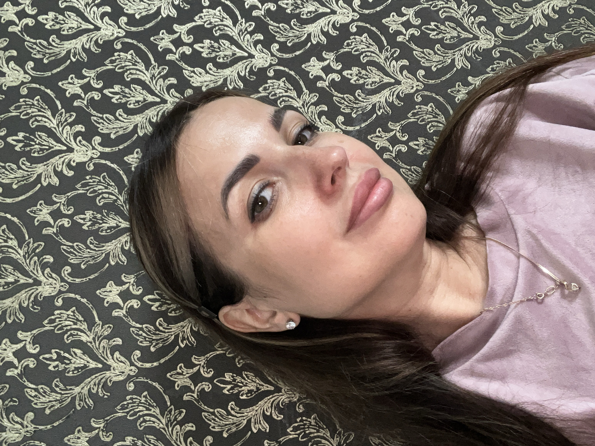 A woman with dark brown hair and brown eyes is positioned in the center of the frame, looking towards the left. She is wearing a pale pink t-shirt and a necklace with a heart-shaped pendant. Her left ear is visible, adorned with a stud earring. The background is a patterned wallpaper with an intricate floral design in gold and dark gray. The lighting suggests an indoor setting, possibly a home. The scene is static, with no apparent action taking place.