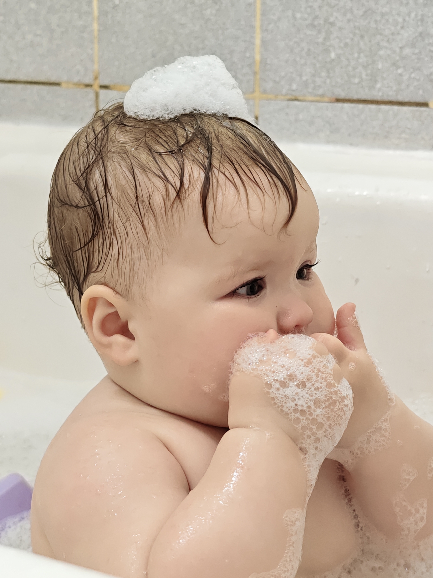 A close-up, eye-level shot captures a baby or young toddler enjoying a bath indoors in Chaikovsky, Russia. The infant, with fair skin and dark, wet hair, is the central subject. A generous pile of white soap bubbles rests on top of its head, indicating a recent hair wash. More frothy, white bubbles cling to the baby's hands, which are brought up towards its mouth, suggesting playful interaction or curiosity with the suds. The baby's dark eyes gaze intently towards the right, out of frame, displaying a calm and engaged expression.
The setting is a white bathtub, with its rim visible in the foreground and lower mid-ground. The background features light-colored, possibly grey or beige, bathroom tiles with darker grout lines, characteristic of a household bathroom. Hints of water droplets and additional bubbles are visible on the baby's skin and around the tub. A small, partially submerged purple plastic object or toy can be seen in the bottom left corner, adding to the bath time ambiance. The lighting is soft and even, highlighting the textures of the bubbles and the baby's features. No text is visible in the image. The overall impression is one of an adorable, contemplative, and playful moment during a baby's bath time.