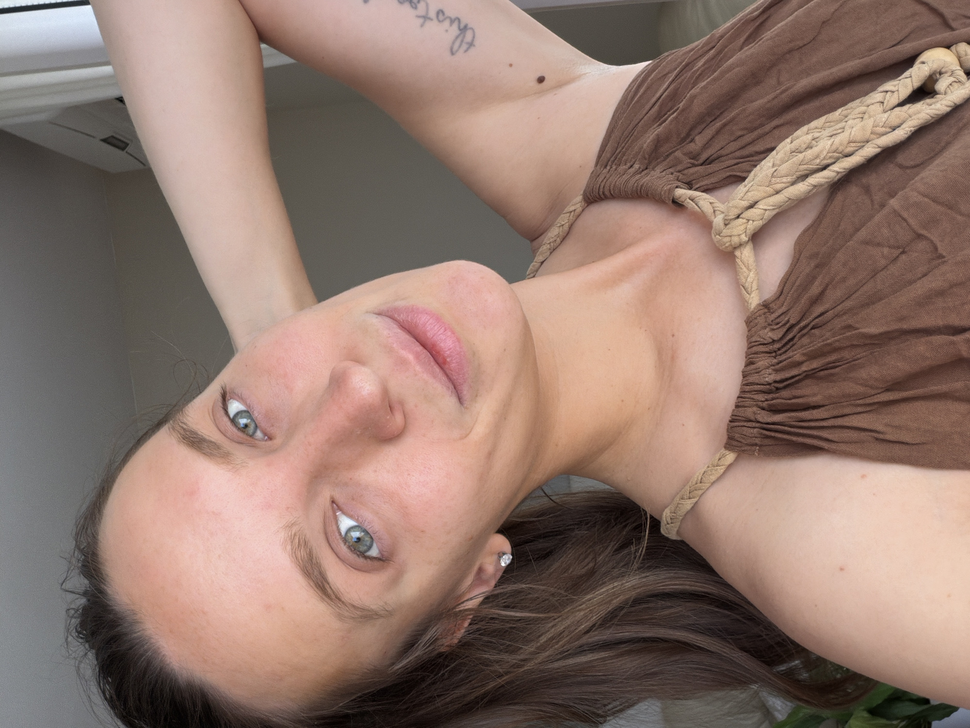 A woman is visible in a close-up shot. The background is a room setting with a window. The woman is wearing a brown dress with braided straps. She has fair skin, light eyes, and brown hair. A tattoo is visible on her arm. The overall image is taken from a low angle. The location of Budva, Montenegro, cannot be determined from the image.