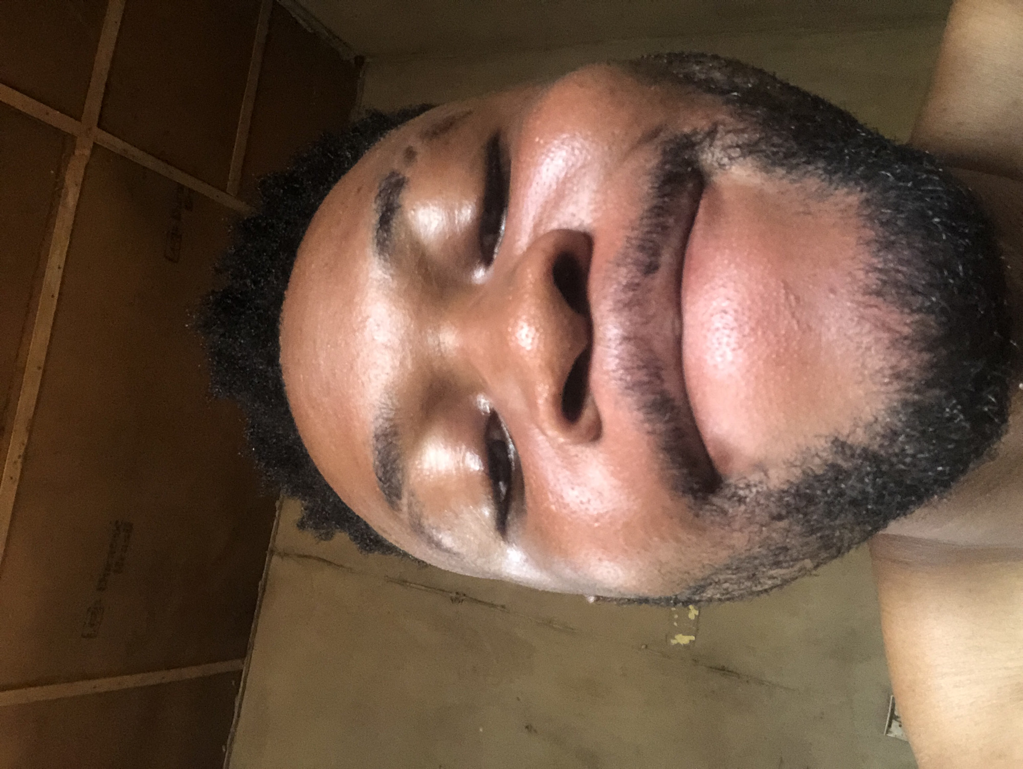 A close-up portrait depicts a dark-skinned person with closed eyes. The person has short, dark hair and a short, dark beard. The skin appears glossy. The background consists of walls with visible wooden support beams. The location is unknown.