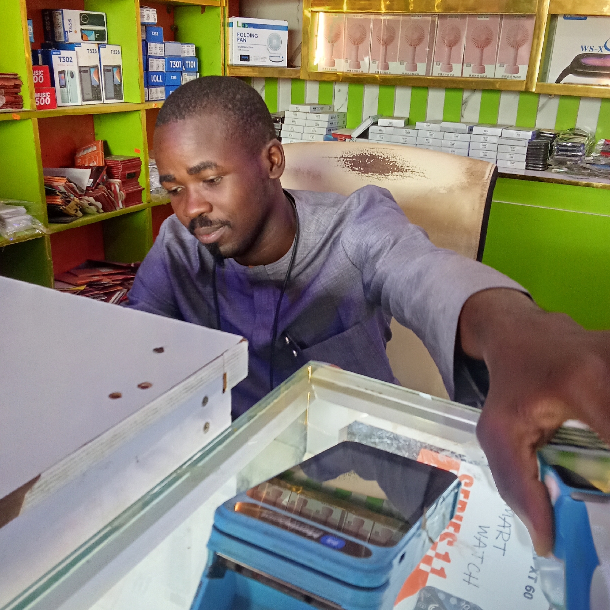 The image depicts a man in what appears to be a retail shop in Akwanga, Nigeria. The man, with short black hair and a mustache, is wearing a gray shirt. He is standing behind a glass counter, reaching for a product, possibly a "SMART WATCH XT 60" as indicated on a nearby box.
Behind the man, there is a shelf filled with various items, including packaged mobile phones labeled with brand names like "TECN0 T302," "T528," "T301," and "T101." Other items on the shelf include packaged portable fans and other electronics. The shop's interior has green and white striped walls.
