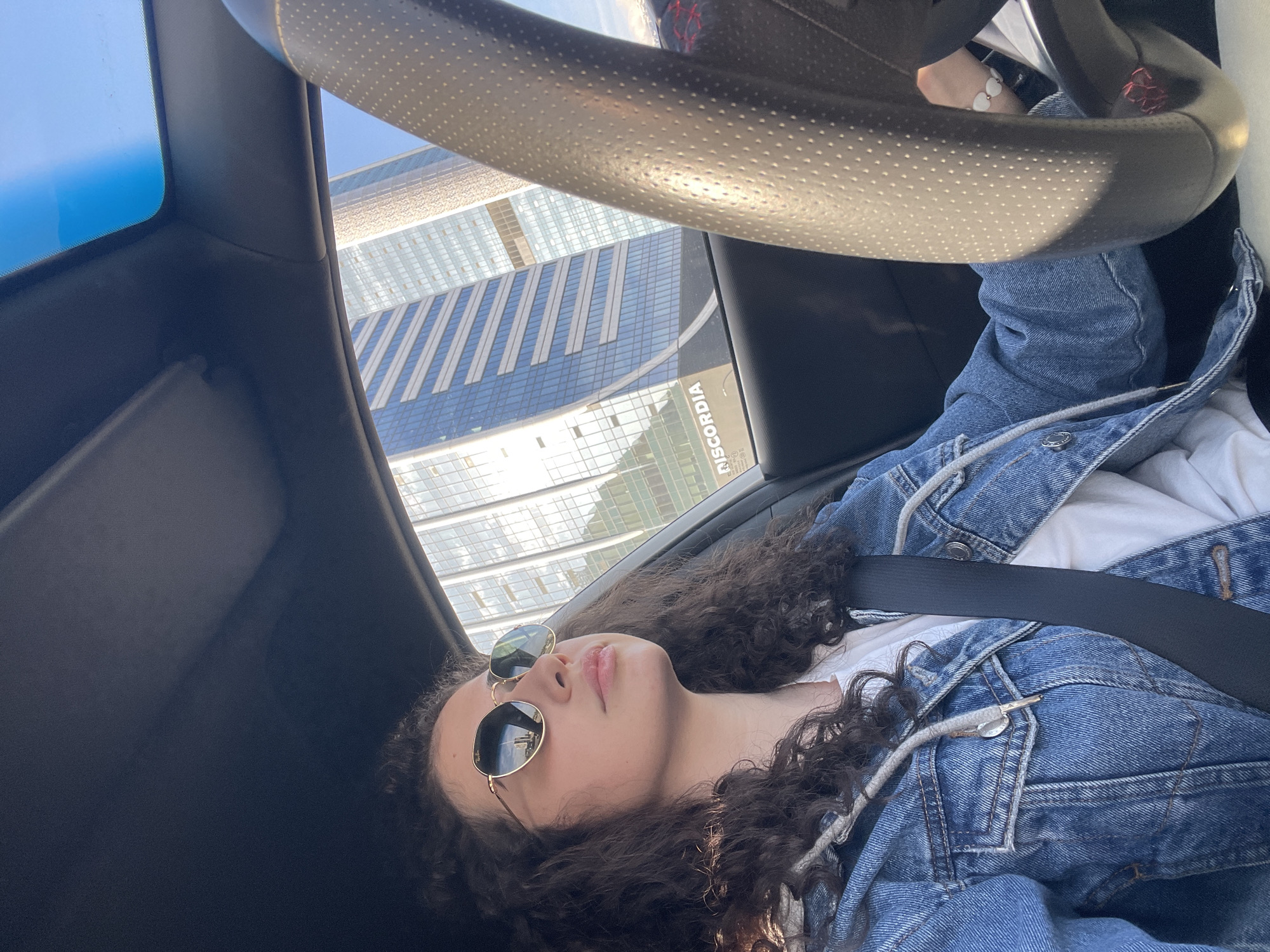 A photograph taken inside a car, potentially a self-portrait, shows a person with curly brown hair, wearing sunglasses and a denim jacket over a white shirt, secured with a seatbelt. The person is reclining in the driver's seat. The steering wheel is visible in the foreground, partially obscuring the view. Through the car windows, a high-rise building with a repeating pattern of glass panels is seen. A sign that reads "ISCORDIA" is visible on the building. Based on the architecture and the building's signage, the location is likely the Capital Fort building in Sofia, Bulgaria. The sky appears to be clear and sunny.