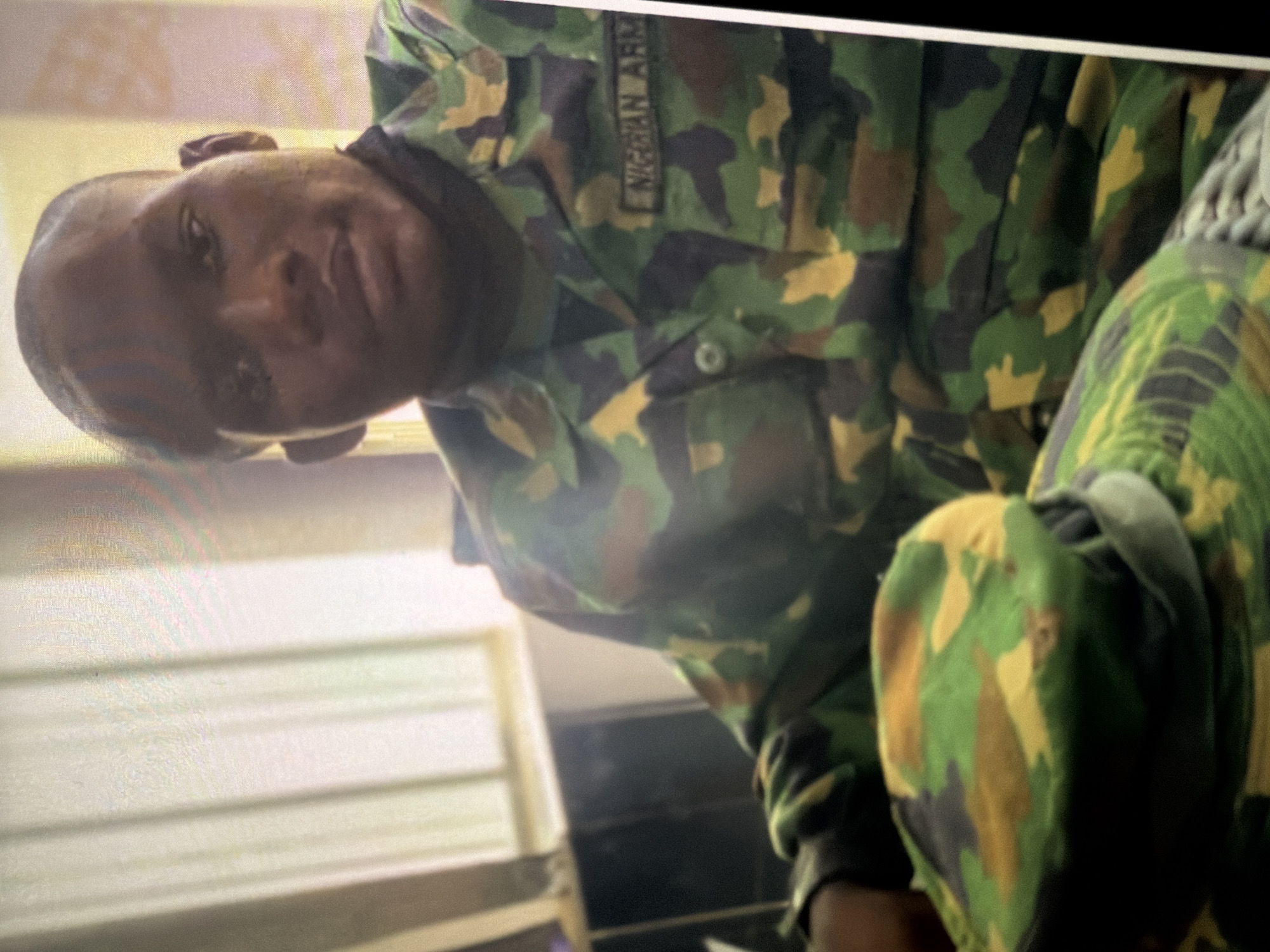 A medium shot photograph shows a young Black male wearing a camouflage military uniform. He is seated, with his head slightly tilted to his left. The uniform jacket is a digital camouflage pattern of greens, browns, and yellows. A name tape on the right side of his chest reads "NIGERIAN ARMY". In the foreground, a camouflage military cap is placed on his lap. Behind him, a window with vertical blinds is visible, with light filtering through. A dark object, possibly a desk or cabinet, is located to his left. The overall lighting suggests an indoor setting. The background is slightly out of focus.