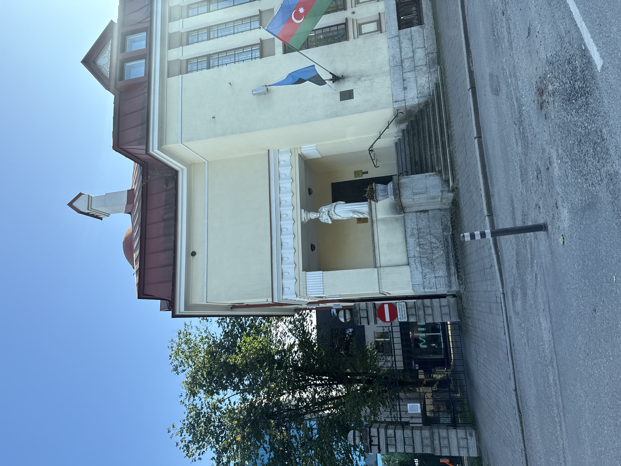 The image shows the exterior of a building in Tallinn, Estonia. The building is light yellow with a red roof and features classical architectural elements, including columns and a pediment. Two flags are flying from the building: the flag of Estonia and the flag of Azerbaijan. In front of the building, there is a set of stairs leading to an entrance with a statue of a woman. To the left of the building, there is a tall green tree and a modern glass-fronted building behind a fence. A street is in the foreground, with a gray asphalt surface and a paved sidewalk. A black and white striped bollard is visible on the sidewalk. There is a sign on the fence with Estonian text. The sky is clear and blue.