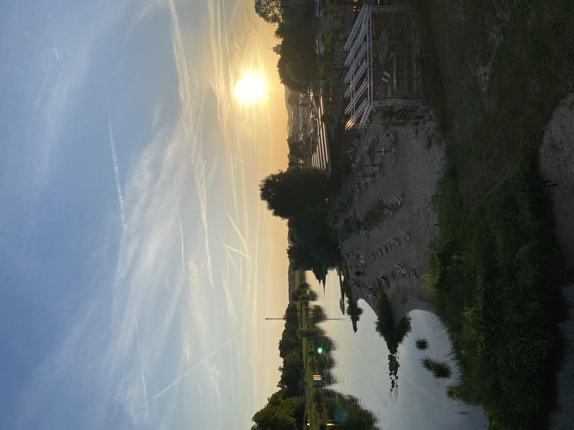 This image depicts a sunset over the Weser river in Rinteln, Germany. On the left side of the frame, a wide, calm river reflects the sky and surrounding greenery. A small, illuminated structure sits on the riverbank. A red cellular tower is visible in the distance. On the right side of the image, a sandy beach area is set up with numerous rows of empty lounge chairs and umbrellas. A small stage or bar structure with a thatched roof is situated on the sand. Several palm trees are visible. A few individuals are scattered on the beach, some sitting in chairs and others walking. The sky is a gradient of blue and orange, with numerous contrails from airplanes streaking across it. The overall scene suggests a relaxed, late-afternoon or early-evening atmosphere at a riverside leisure area.