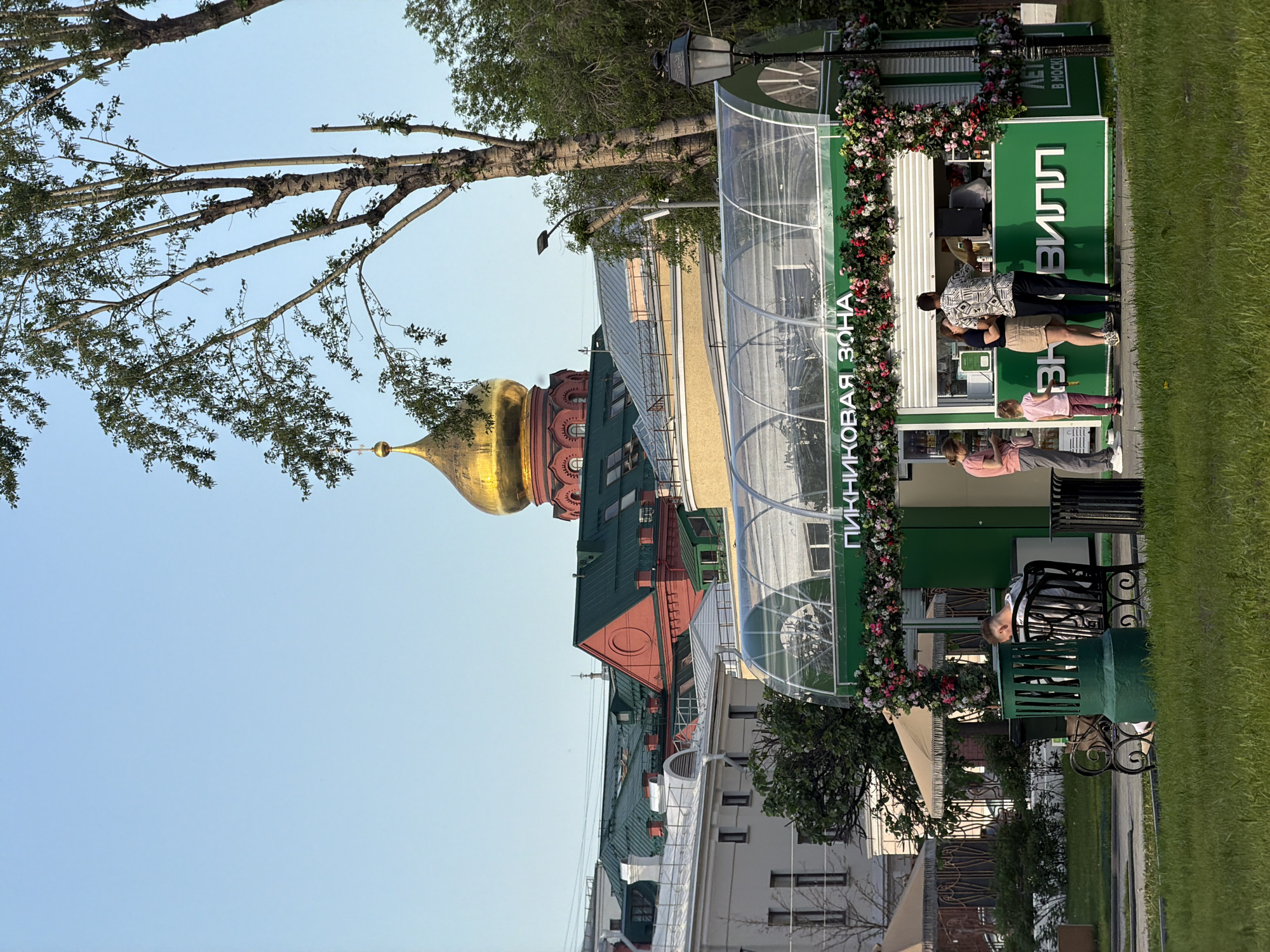 The image captures an urban scene in Moscow, Russia, with a mix of architectural landmarks and everyday life. In the background, a prominent golden dome of a Russian Orthodox church rises above the rooftops of buildings, suggesting the city's rich religious heritage.
The foreground features an outdoor "picnic zone," marked by signage in Russian. Several people are present: a woman is sitting on a bench, and a group consisting of two women, one man and one child are standing in front of what appears to be a booth or service station within the picnic area. The people are dressed in casual clothing. The picnic area is adorned with flowers, adding a touch of nature to the urban landscape.
A tree with leaves frames the left side of the image, contrasting with the buildings and urban elements. The sky is clear and blue, providing a bright backdrop for the scene.