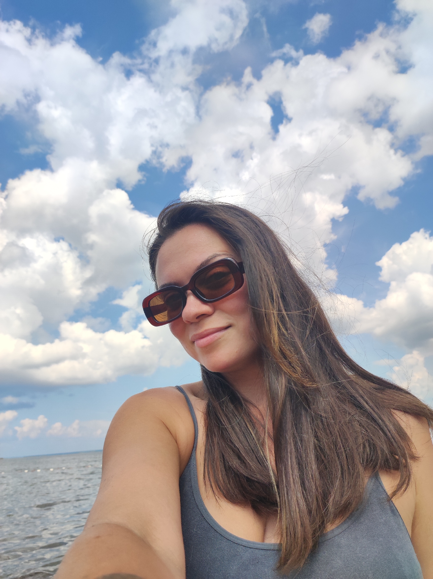 A woman with long, dark hair and sunglasses is taking a selfie outdoors. She is wearing a gray tank top. The background features a bright blue sky with white, fluffy clouds and a body of water, likely the Kama River, which is a significant feature of Chaikovsky, Russia. The scene appears to be a sunny day, possibly at a beach or waterfront area within the city.