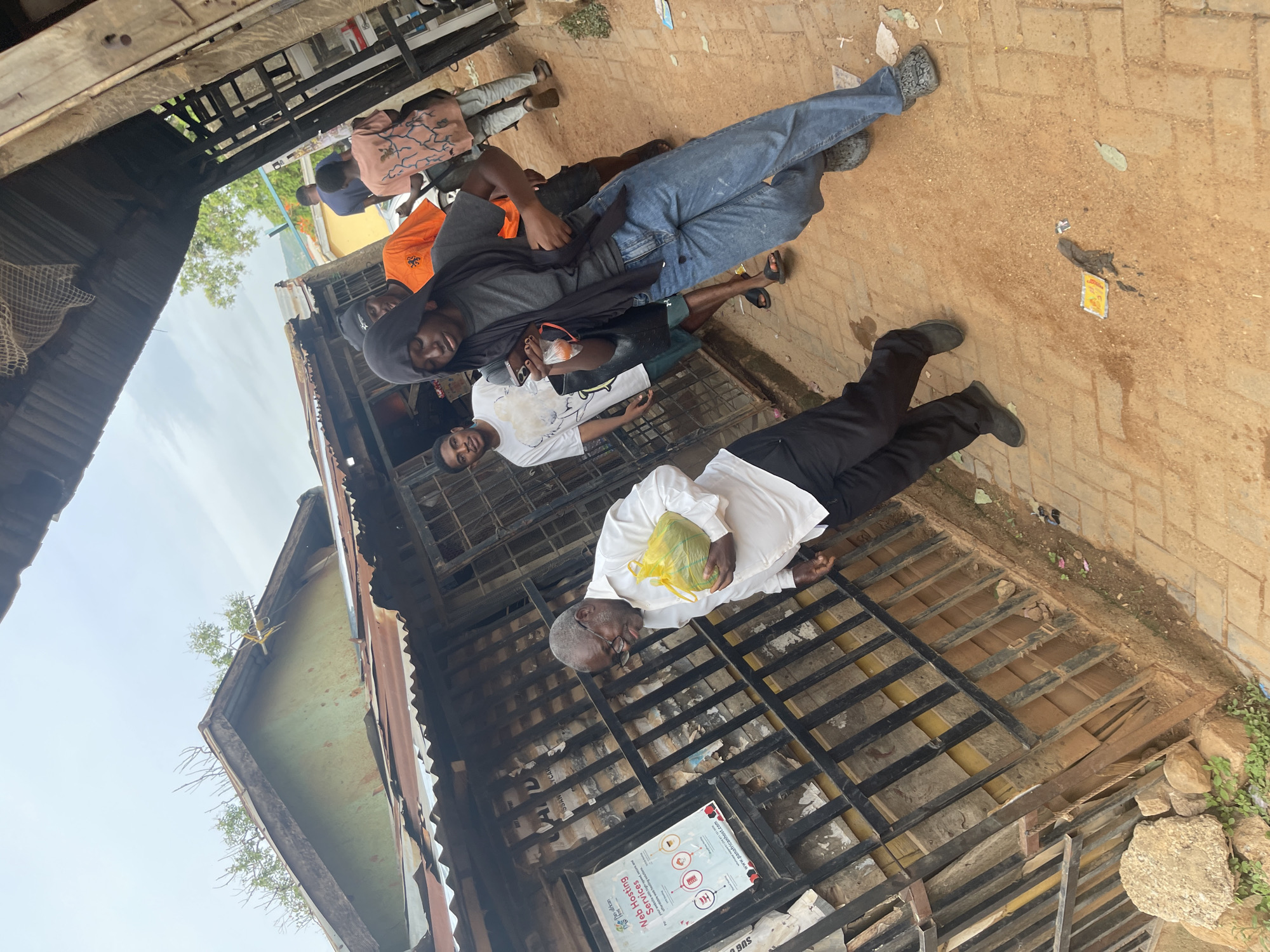 The image depicts a street scene in Jos, Nigeria, with several people interacting outdoors. In the foreground, an older man wearing glasses, a white shirt, and dark pants is lying down, holding a yellow bag. He appears to be resting or perhaps unwell.
Behind him, several younger individuals are visible, some standing and others seated. One person in blue jeans and a dark shirt is leaning against a structure. Another person in an orange t-shirt is also present. The overall setting appears to be a somewhat informal, perhaps market or roadside area, with metal grates and corrugated metal roofing visible. The ground is a mix of dirt and paved bricks.
The lighting suggests it is daytime, likely outdoors with natural light. The weather appears to be fair, with a bright sky visible in some parts of the image. There is some clutter and debris on the ground, typical of a busy outdoor environment.
A poster with text is partially visible on a metal grate, with the words "Web Hosting Services" and some icons indicating services. Other small pieces of paper and debris are scattered around. The general atmosphere seems casual, with people gathered in what could be a public space. The interactions between the individuals are not clearly defined, but there's a sense of community or shared space. The image captures a slice of everyday life in this Nigerian location.