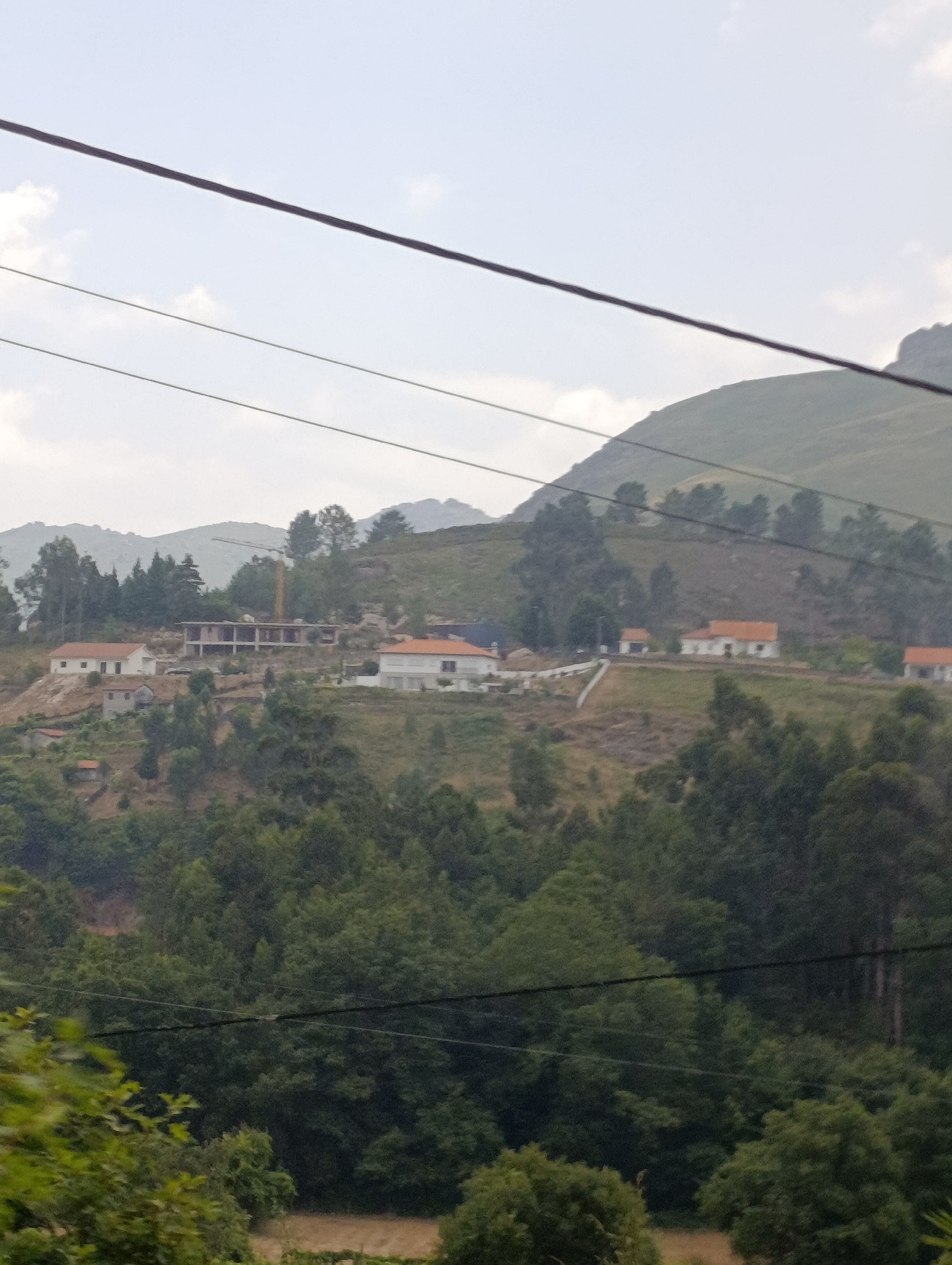 The image displays a hillside landscape in Terras de Bouro, Portugal. Several houses with orange-tiled roofs are scattered across the incline, interspersed with patches of dense green foliage and trees. A construction crane is visible on the upper part of the hill, suggesting ongoing building activity. Power lines stretch across the frame, dividing the sky from the landscape. The overall scene is one of rural development on a hilly terrain under a cloudy sky.
