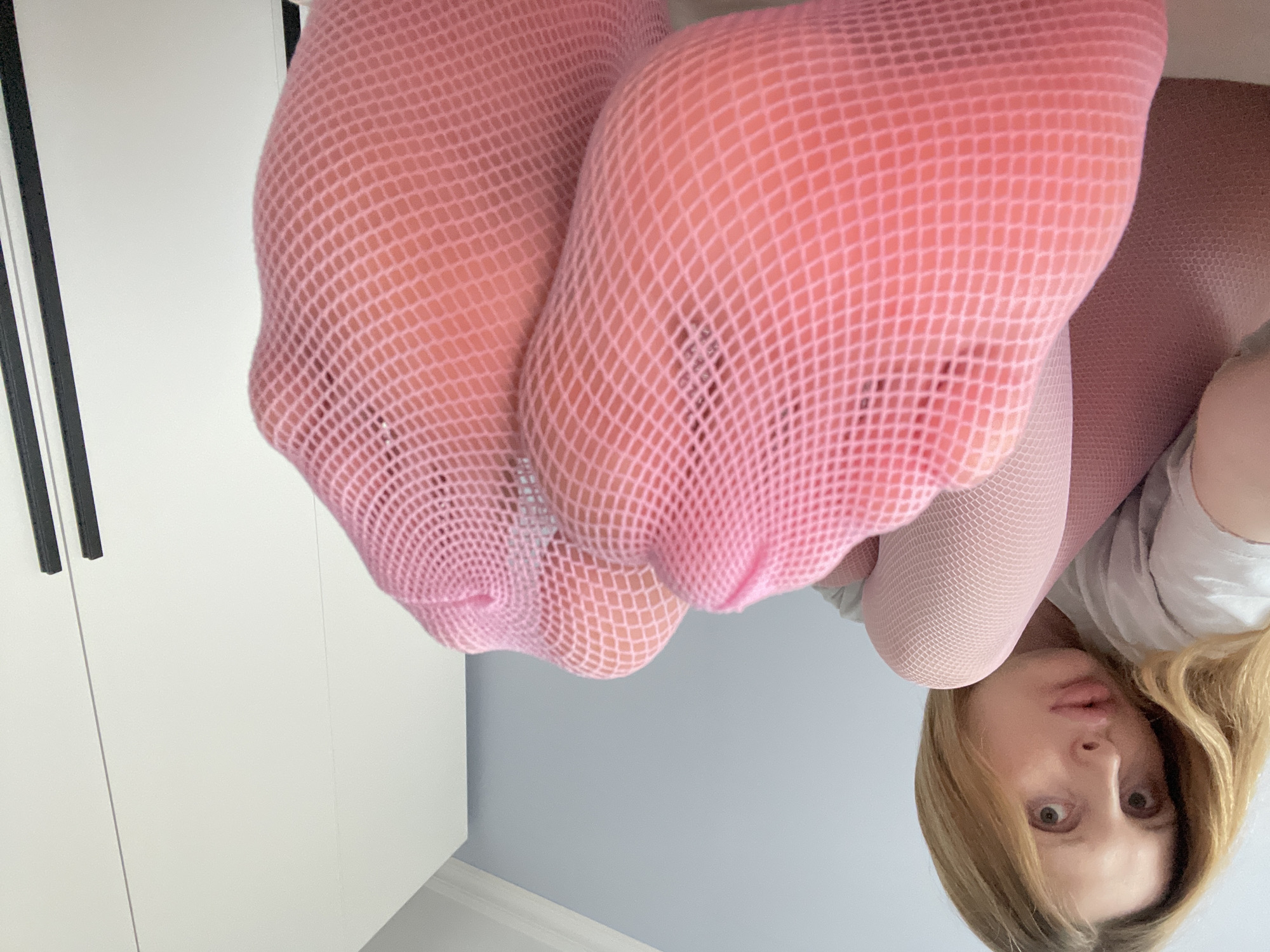 The media file shows a low-angle, upside-down shot of a person's legs and face. The subjects are a person wearing pink fishnet tights. Their legs are prominently featured in the foreground, with the mesh pattern of the tights clearly visible. The person's face is in the lower right portion of the image, with wide eyes and slightly parted lips, appearing to be looking directly at the camera.
The setting appears to be indoors, with a white wardrobe visible on the left side of the image. The background consists of a light blue wall with a white baseboard. The lighting suggests it is daytime. There is no visible text in the image. The overall mood is somewhat playful or suggestive due to the pose and attire. The location context is Yablonovskiy, Russia.