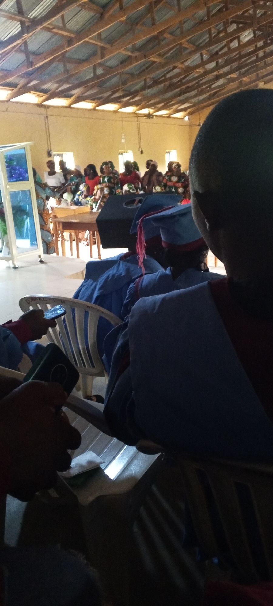 The image captures an indoor gathering, possibly a church or community event, in Bahit, Nigeria. In the foreground, a person is partially visible from the back, wearing a blue graduation gown and cap with a pink tassel. Their left hand is holding a smartphone, and their right hand holds a dark object. To the left, another person's hands are visible, one holding a smartphone and another a green object. A white chair with a curved back is situated between them.
In the mid-ground, several individuals are seated or standing, many wearing colorful attire and headwraps. A wooden table is positioned in front of them. Behind the table, a large black speaker or podium is visible. A blue and white podium or display stand is also on the left side of the mid-ground.
The environment appears to be a room with a wooden beamed ceiling and light-colored walls. Several windows are visible in the background, allowing natural light to enter the space. The overall impression is that of a formal occasion taking place within a community hall or similar venue.