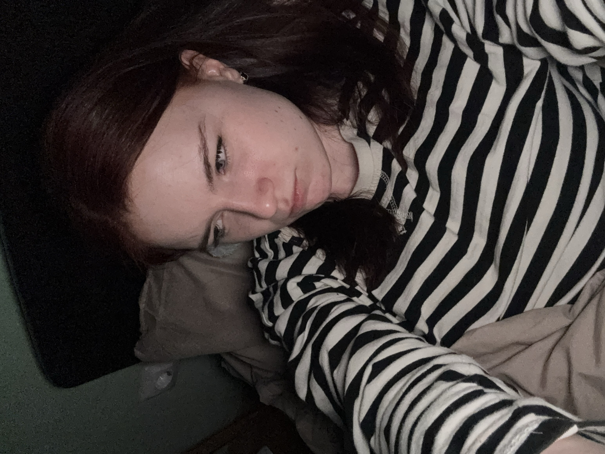A young woman with auburn hair lies on a bed or couch, her head resting on a light brown pillow. She wears a black and white striped long-sleeved shirt or pajamas. The woman's face is partially visible, showing a melancholic expression. The background is dark, suggesting a dimly lit room with a dark headboard or wall behind her. A portion of the wall is green in the bottom left corner. The setting appears to be indoors, likely a bedroom. There is no evidence to suggest that the photograph was taken in St. Petersburg, Russia.