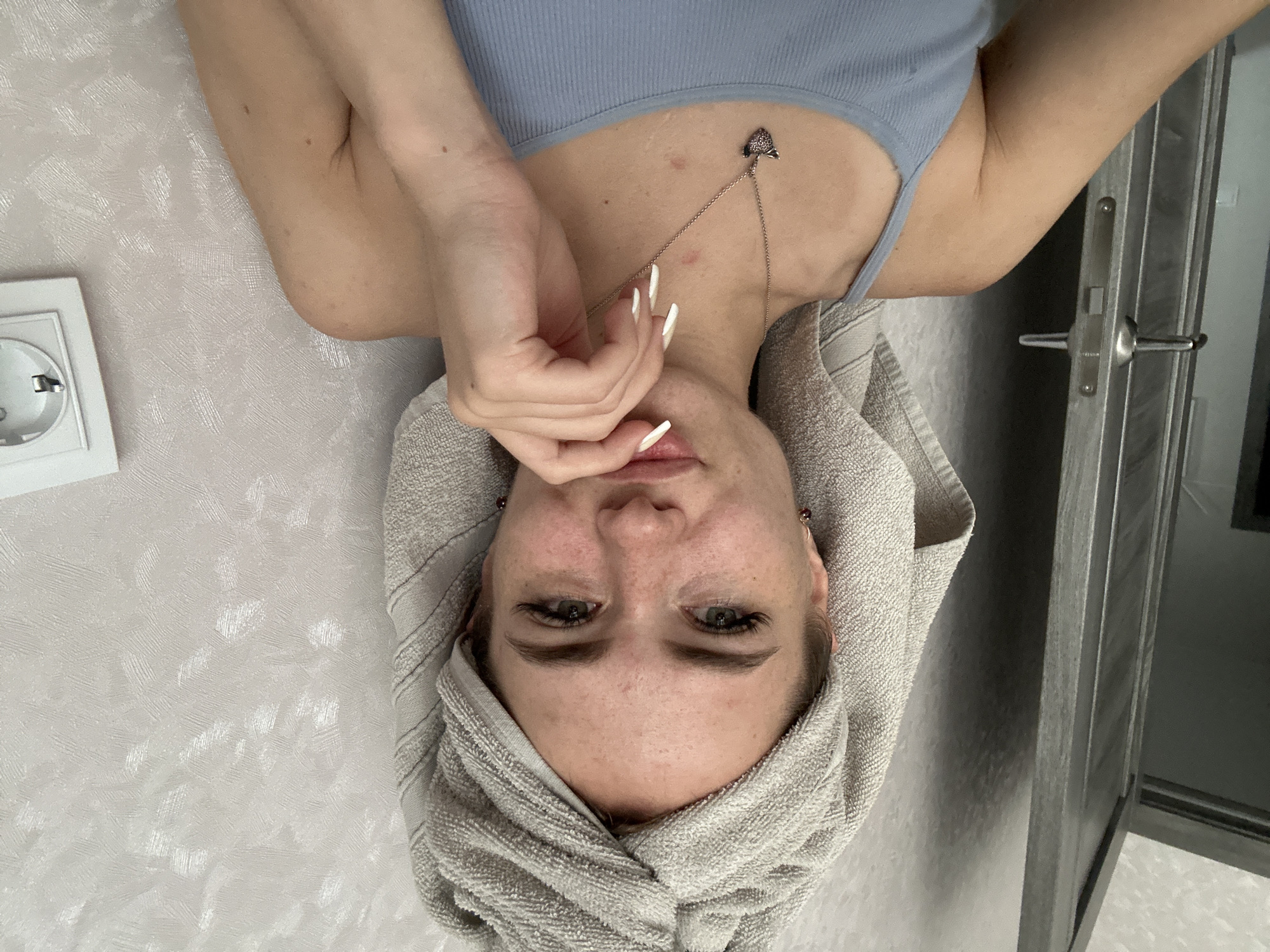 A young woman with a grey towel wrapped around her head is looking at the camera. She has green eyes, fair skin with freckles, and is wearing a light blue tank top and a silver necklace with a heart-shaped pendant. Her right hand is resting on her chin. The background is a textured, light-colored wall. To the left of the woman is a closet door with a silver handle. To the right, a white electrical outlet is visible on the wall. The scene appears to be indoors, likely in a residential setting.