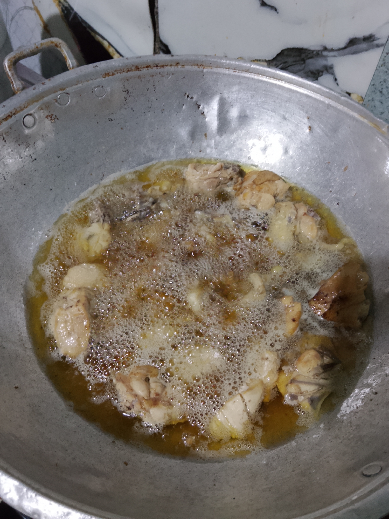A close-up, overhead shot shows pieces of chicken being deep-fried in a large, well-used aluminum wok. The oil is bubbling vigorously around the chicken pieces, which appear to be light brown and golden. The wok is situated on a surface that appears to be a countertop with a marbled pattern, possibly in a kitchen. The background is out of focus, but hints of a tiled wall and possibly other cooking utensils can be seen. The image conveys the action of cooking and the sensory experience of frying food, suggesting a domestic or commercial kitchen setting. There is no information about the city of Merbopuntong, Indonesia in the image or the provided crops.