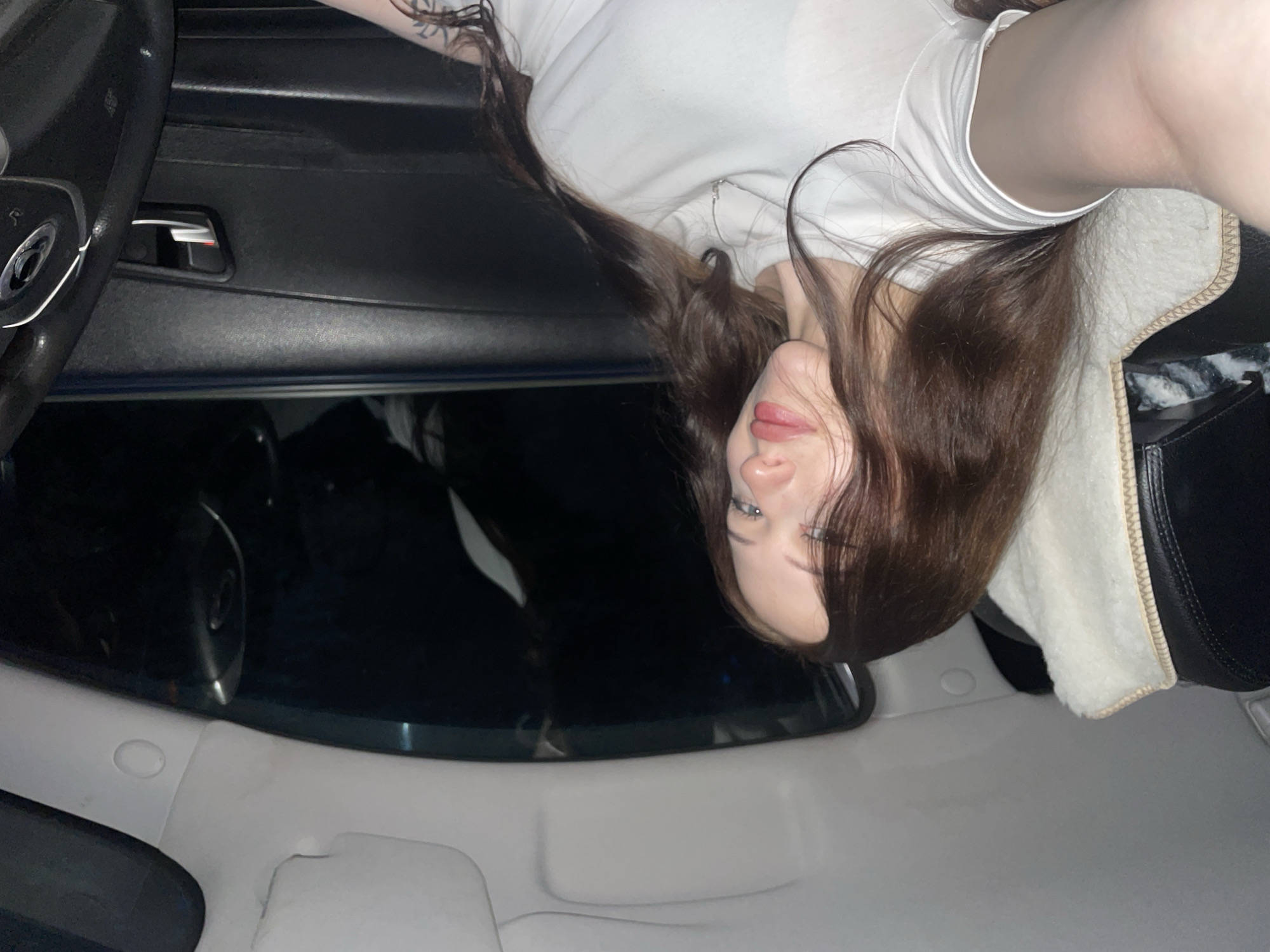 The image shows a person lying down inside a vehicle, possibly a car. The person is wearing a white t-shirt and has long, dark brown hair. They are looking towards the camera with their eyes open. The interior of the vehicle is dimly lit, with parts of the dashboard and seats visible. The background is dark and out of focus, suggesting the vehicle may be parked in an outdoor location at night. There is no discernible information about the specific location within the city of Gavrilovka, Russia, or any other identifying details about the city.