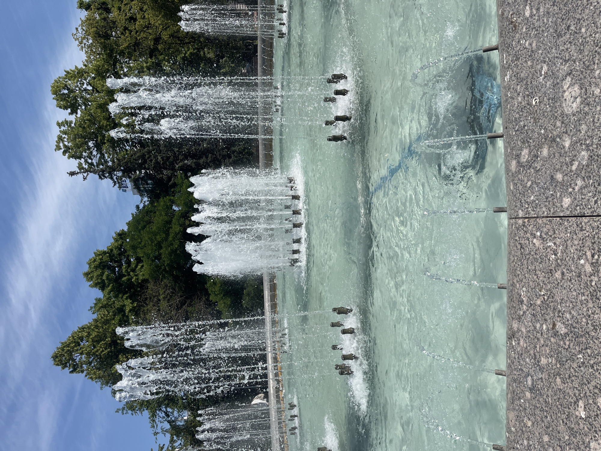 The image depicts a large, multi-tiered fountain in a city park, likely in Krasnodar, Russia. The fountain features numerous jets of water spraying upwards, creating a dynamic and refreshing visual. The water itself is a clear, turquoise hue, and the pool is shallow, allowing glimpses of the submerged fountain mechanisms. The foreground is dominated by the edge of the fountain basin, constructed from a speckled grey granite. Behind the fountain, a lush backdrop of green trees forms a natural wall, with the blue sky and scattered white clouds overhead. In the distance, a tall building with the logo "BTB" is visible, suggesting an urban setting. There are no people visible in the image.