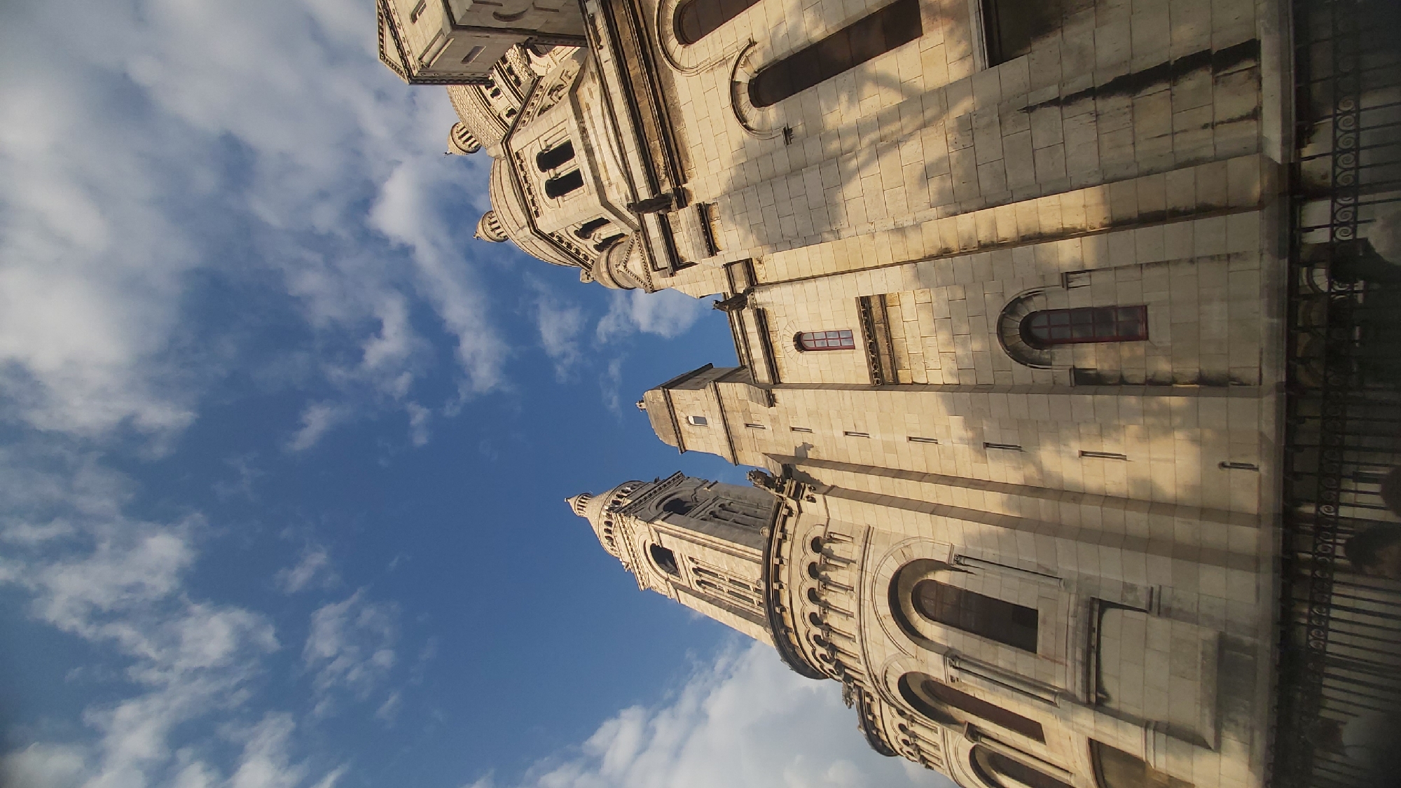 The image shows a low-angle view of the Sacré-Cœur Basilica in Montmartre, Paris, France. The basilica is constructed of light-colored stone and features multiple domes and a tall bell tower. The architecture is Romanesque-Byzantine, characterized by rounded arches and intricate detailing. The sky above is a vibrant blue, dotted with white, wispy clouds. Sunlight illuminates the facade of the basilica, casting long shadows that accentuate its architectural features. In the foreground, a black wrought-iron fence is visible, with the tops of a few people's heads just visible behind it, suggesting they are looking up at the basilica. The overall impression is one of grandeur and architectural beauty against a bright sky.