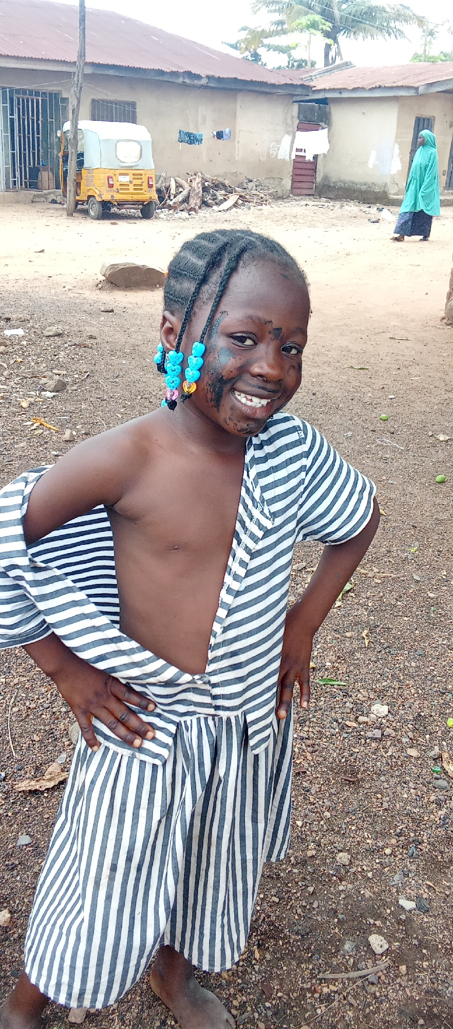 The image depicts a young girl with braids adorned with colorful beads, standing outdoors on what appears to be a dirt or gravel ground. She is wearing a striped garment, partially open, revealing her chest. Her face has smudges of a dark substance, possibly dirt or paint. She is smiling with her hands on her hips.
In the background, there is a yellow tricycle, a simple building with laundry hanging outside, and a woman wearing a green hijab walking away. The scene appears to be located in Akwanga, Nigeria, based on the visual cues suggesting a rural or semi-urban setting with modest infrastructure.