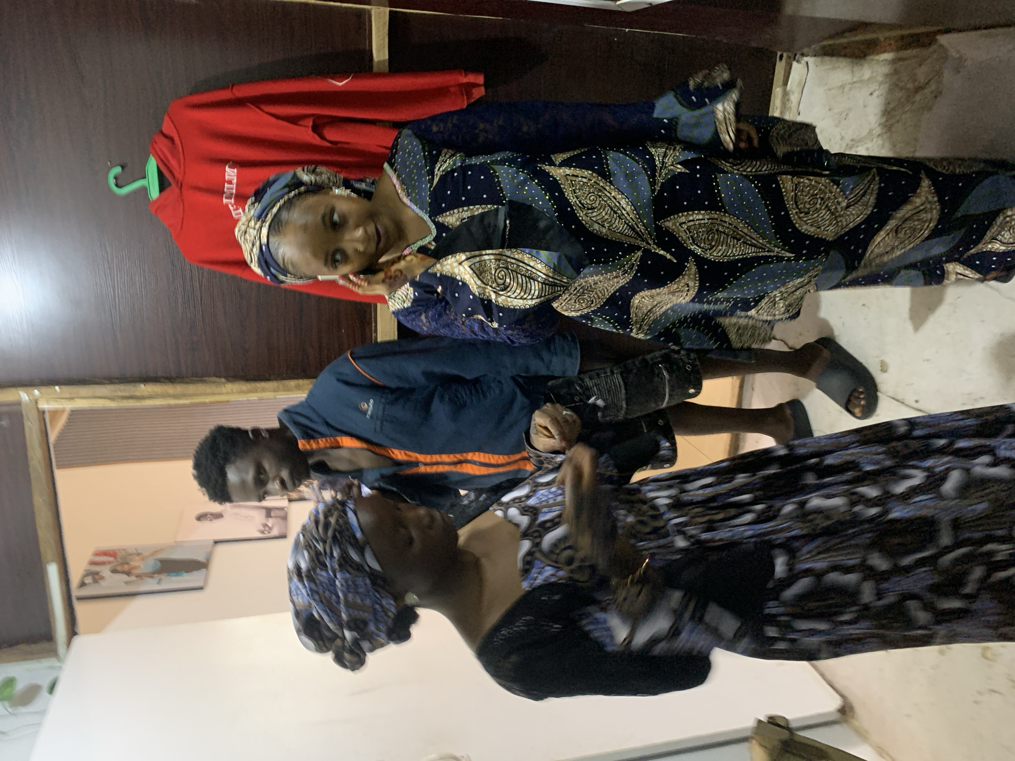 The image shows three people indoors in what appears to be a home in Jos, Nigeria. On the left, a woman in a patterned headscarf and dress is interacting with a man in a blue jacket with orange trim, who is wearing shorts and sandals. To the right of the man, a woman in a patterned dress and headscarf poses with her hand to her face. Behind her is a wooden door on which a red hooded sweatshirt hangs. Pictures hang on the wall. The room appears to be illuminated by artificial light.