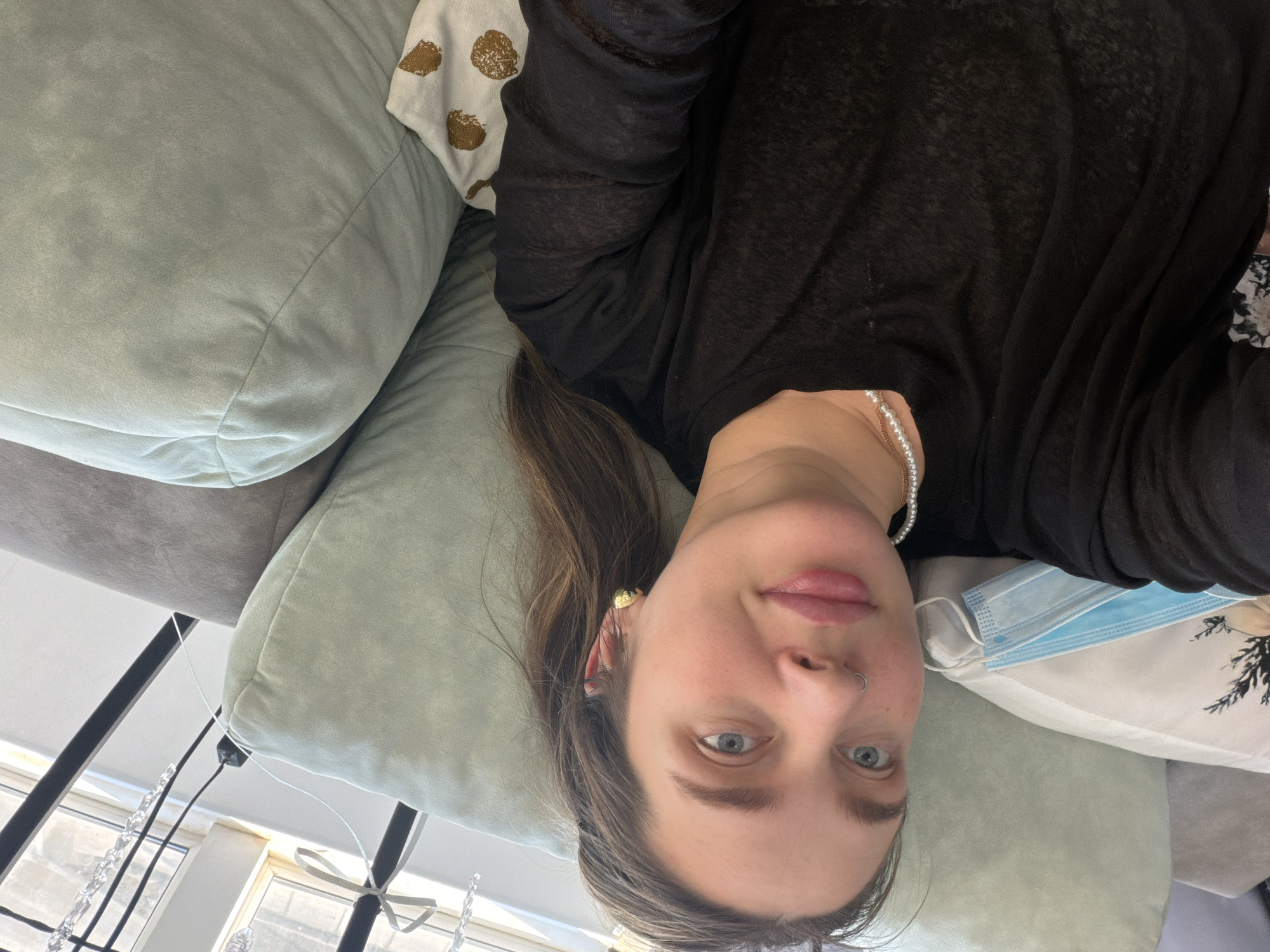 A female individual is depicted reclining on a sofa in an indoor residential setting. She is positioned to face the camera directly, displaying light blue eyes, light pink lips, and light brown hair. Accessories include a silver nose ring in her left nostril, a gold-colored stud earring in her right ear, a pearl necklace, and a thin gold chain. She wears a black, translucent long-sleeved top. To her left, a light blue disposable medical face mask rests on a white pillow adorned with a dark botanical pattern. She is supported by light green, suede-like cushions, and a white throw pillow with gold circular patterns is partially visible on her right. A window is present in the background, showing daylight, in front of which stands a black metal frame decorated with clear crystalline elements and a light-colored ribbon. The visual information present in the image does not provide specific identifiers for a precise location within Chaikovsky, Russia.