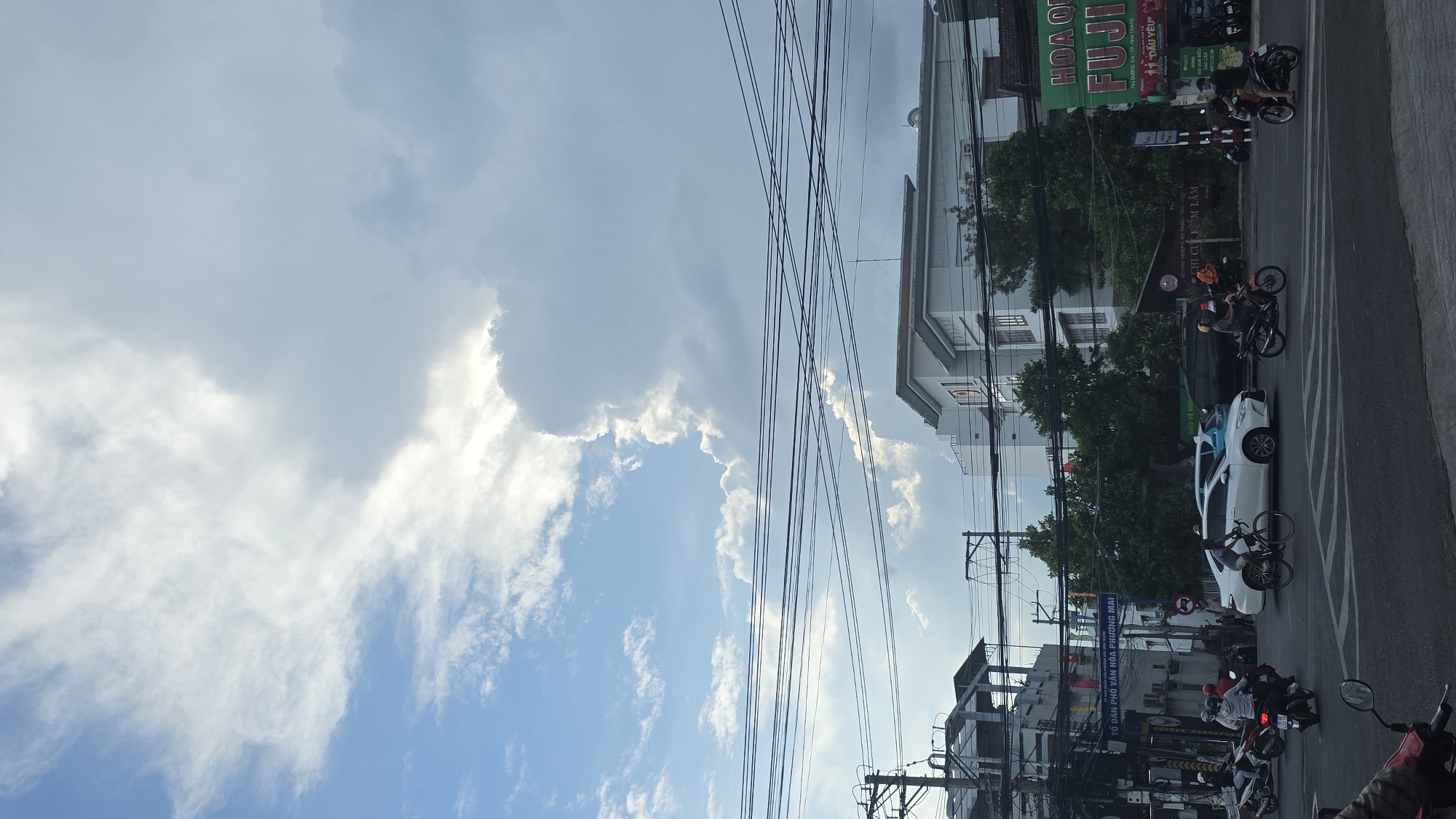 The image captures a street scene in Nha Trang, Vietnam, under a partly cloudy sky. Power lines crisscross the view. Buildings line the right side of the street, including one with a "FUJI" sign. Several motorbikes and a white car are visible on the street.