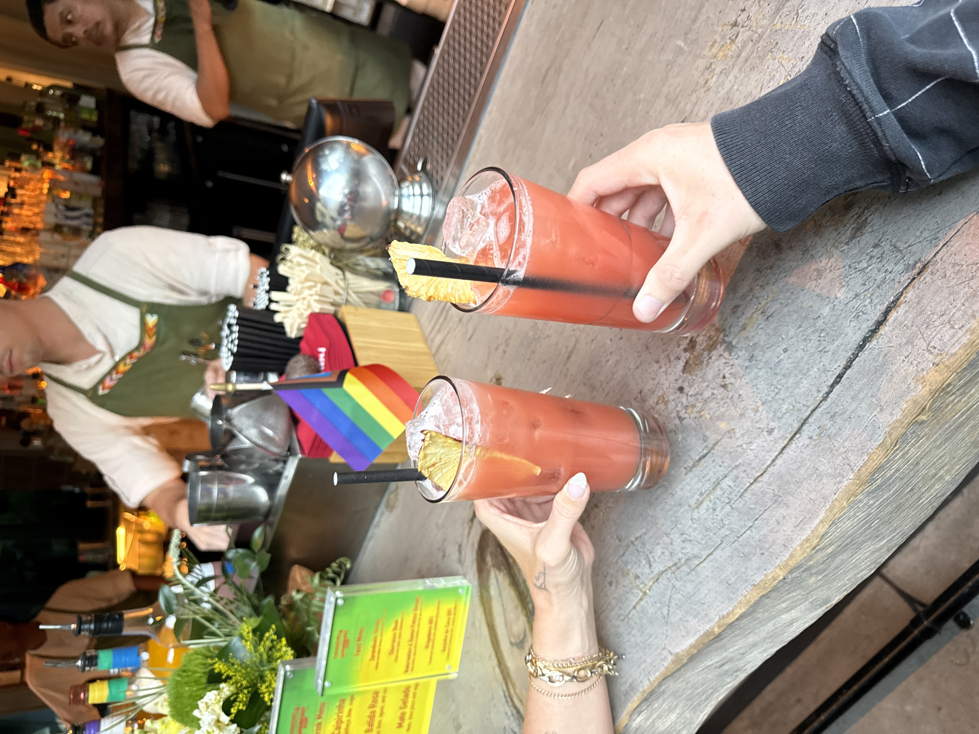 This image captures a close-up view of two hands holding tall glasses filled with a red-colored beverage and ice, garnished with pineapple slices and black straws. The glasses sit on a weathered wooden bar top. In the background, behind a display of bottled drinks and bar equipment, two individuals in aprons are visible, presumably bartenders. To the left, a menu board lists drink names. A small rainbow flag, a symbol of LGBTQ+ pride, is prominently displayed near the drinks. The setting appears to be a bar or restaurant. The image does not provide specific details to determine the exact location within Los Angeles, United States of America.