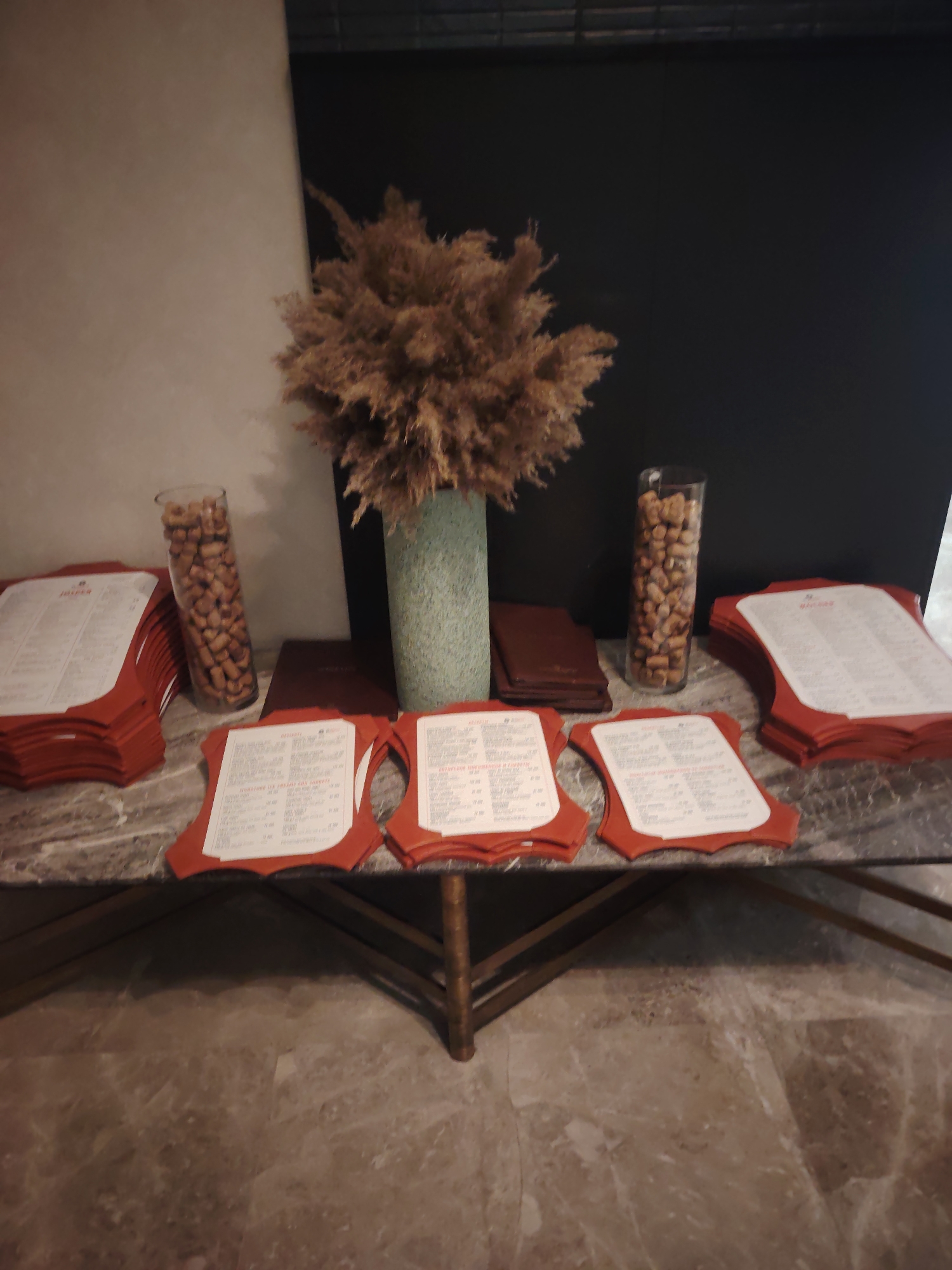 A still life photograph depicts a marble-topped table with several stacks of menus. The menus are red with white pages displaying text and pricing. In the center of the table, a tall, cylindrical vase holds a bundle of dried, feathery foliage. To the left of the foliage, another tall, cylindrical vase is filled with what appear to be nuts or seeds. To the right of the foliage, a dark red, folded booklet rests on the table. The background is divided into a lighter, textured wall on the left and a dark, smooth surface on the right. The table itself is supported by thin, dark metal legs. The floor beneath the table is a polished, marbled surface. The scene is devoid of any people or animals. The overall impression is one of an indoor setting, possibly a restaurant or entryway, prepared for a formal occasion. The location in Konigil, Uzbekistan, is not discernible from the image's visual elements.