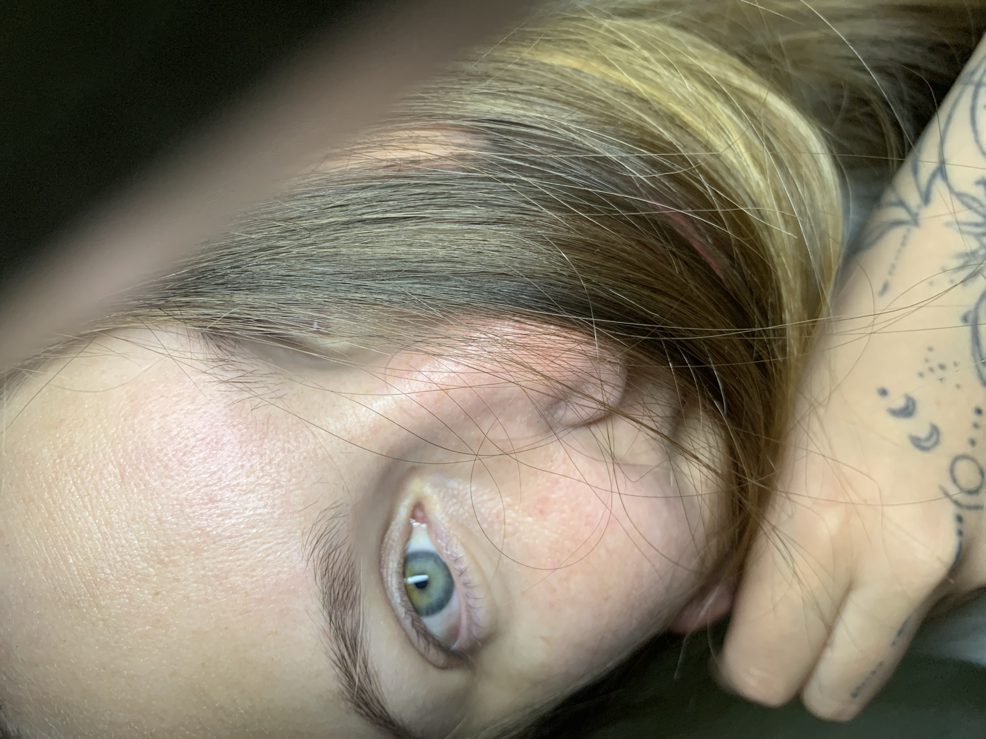 A close-up, angled shot of a person's face and hand, tilted to the side. The person has fair skin, light brown and blonde hair, and green eyes. The hand is holding the side of the head, revealing a tattoo on the hand. The background is dark and out of focus. There is no indication of location and nothing to suggest it is Chaikovsky, Russia.