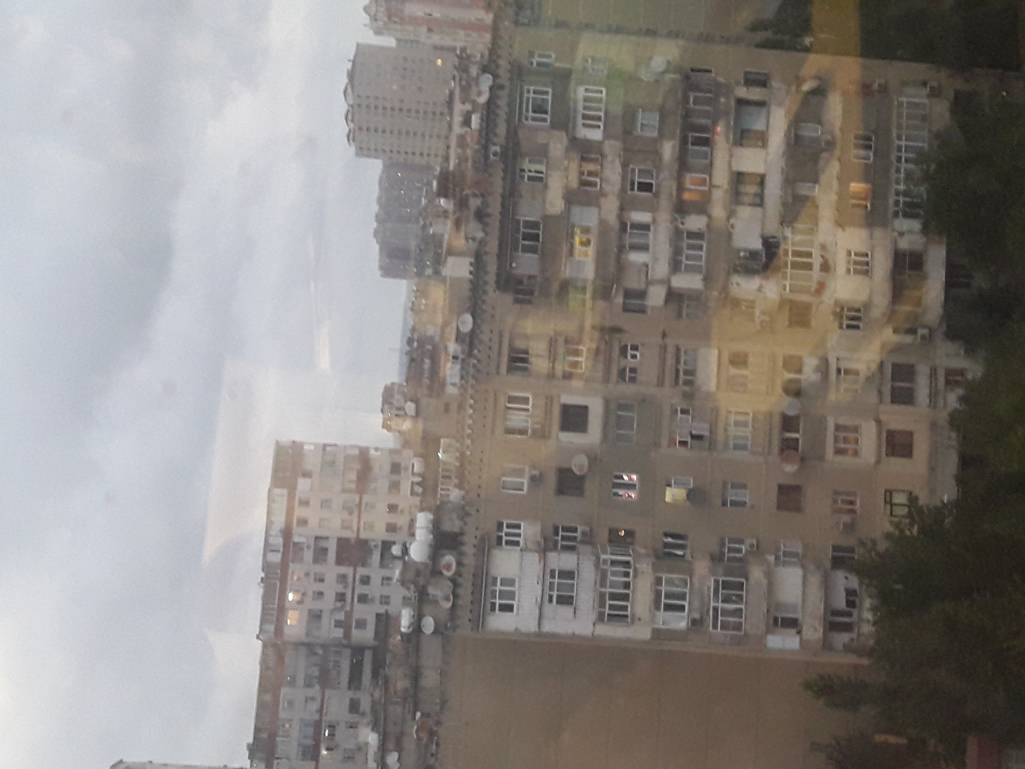 The image is a medium shot of a city skyline on an overcast day. In the foreground, there is a large, multi-story apartment building with a textured, beige facade. The building has numerous windows, many of which appear to be dark or reflecting the cloudy sky. Some windows are lit from within. Balconies with white railings are visible on some floors, and there are satellite dishes mounted on the exterior. Trees with dark green foliage are present at the base of the building. Behind this building, other apartment buildings of varying heights and architectural styles are visible, stretching into the distance. The sky is filled with dense, gray clouds. The overall impression is one of a dense urban environment under a gloomy sky.