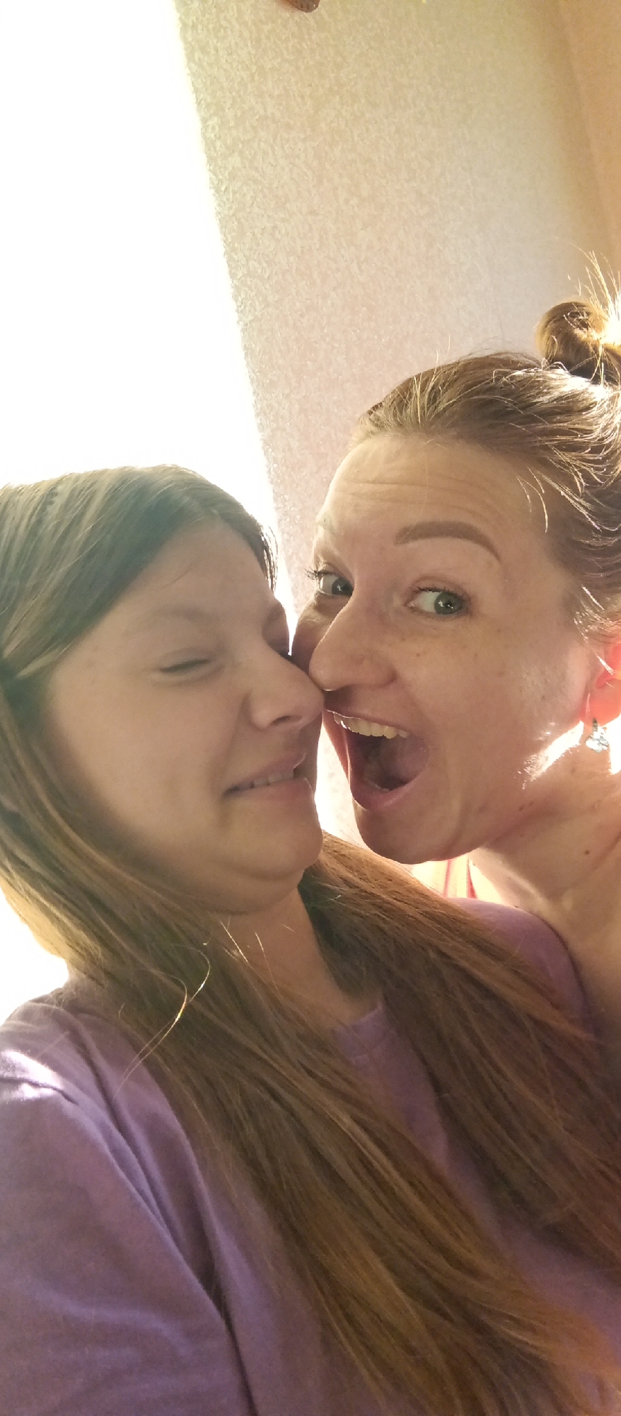 Two females are close to each other, posed for a selfie. The female on the right has her hair in a bun, and her mouth is open wide in a display of excitement. The female on the left is grimacing with one eye closed. The location is Unknown, Unknown. The environment is indoors.