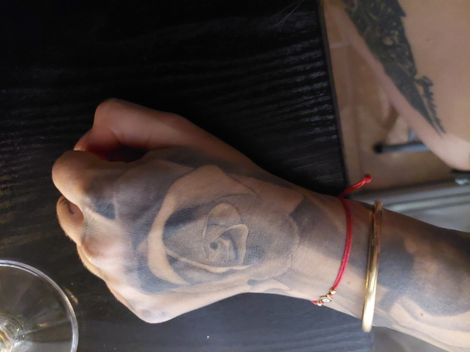 A close-up shot depicts a person's left hand resting on a dark, possibly wooden, surface, likely a table. The hand is heavily tattooed with a rose design that covers a large portion of the back of the hand. The skin tone is light, but obscured by the ink. A thin red string bracelet with small gold beads is worn on the wrist, along with a gold bangle. The hand is slightly clenched. A portion of a glass stemware is visible in the lower left corner of the frame, suggesting a dining or drinking environment. To the right, partially out of focus, is another tattooed arm. Based on the objects depicted, and the tattoos, the location is unknown, but could potentially be in a bar or restaurant in the city of Plovdiv, Bulgaria. More context would be needed to identify the exact location.