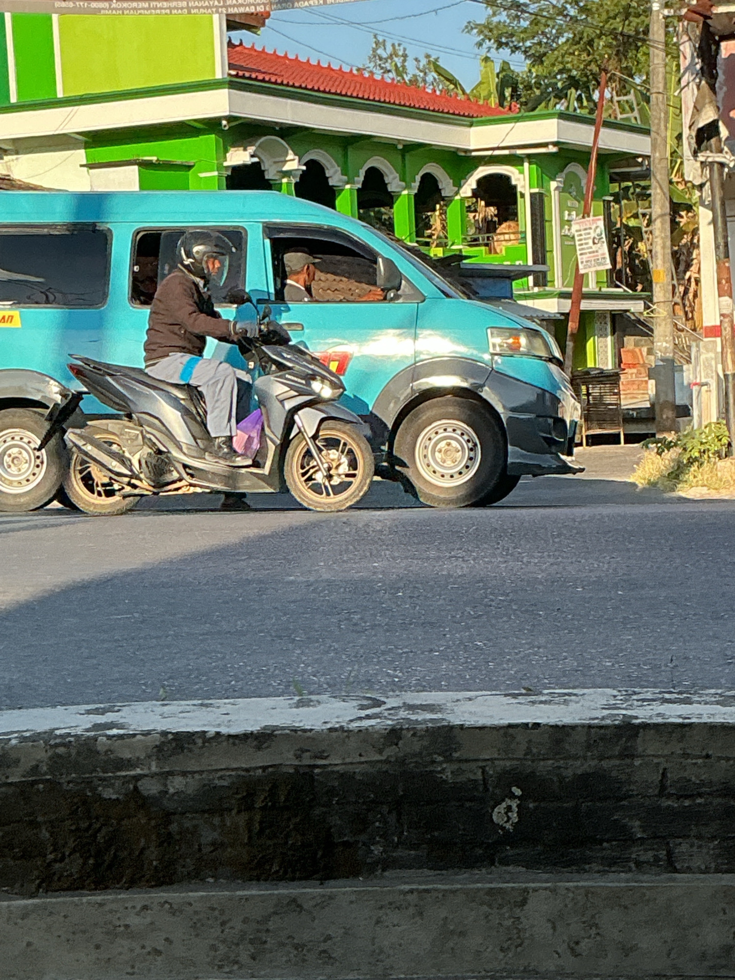 A medium shot captures a motorbike rider wearing a black helmet and a dark jacket with light gray pants, riding a dark-colored scooter on an asphalt road. The rider is positioned on the right side of the frame, slightly angled towards the left, with their left leg extended forward. A purple plastic bag is visible on the floorboard of the scooter. Behind the scooter, a teal-colored van is visible. The van has a black bumper and a black fender flares around its wheels. Its front left wheel is prominent in the foreground, showing a silver rim. In the background, a building with a green facade and a red roof is seen. The building has white arched accents and is partially obscured by trees. A utility pole with wires is also visible to the right of the building. The scene is lit by natural daylight, casting shadows on the road. The location is identified as Kebonrejo, Indonesia.