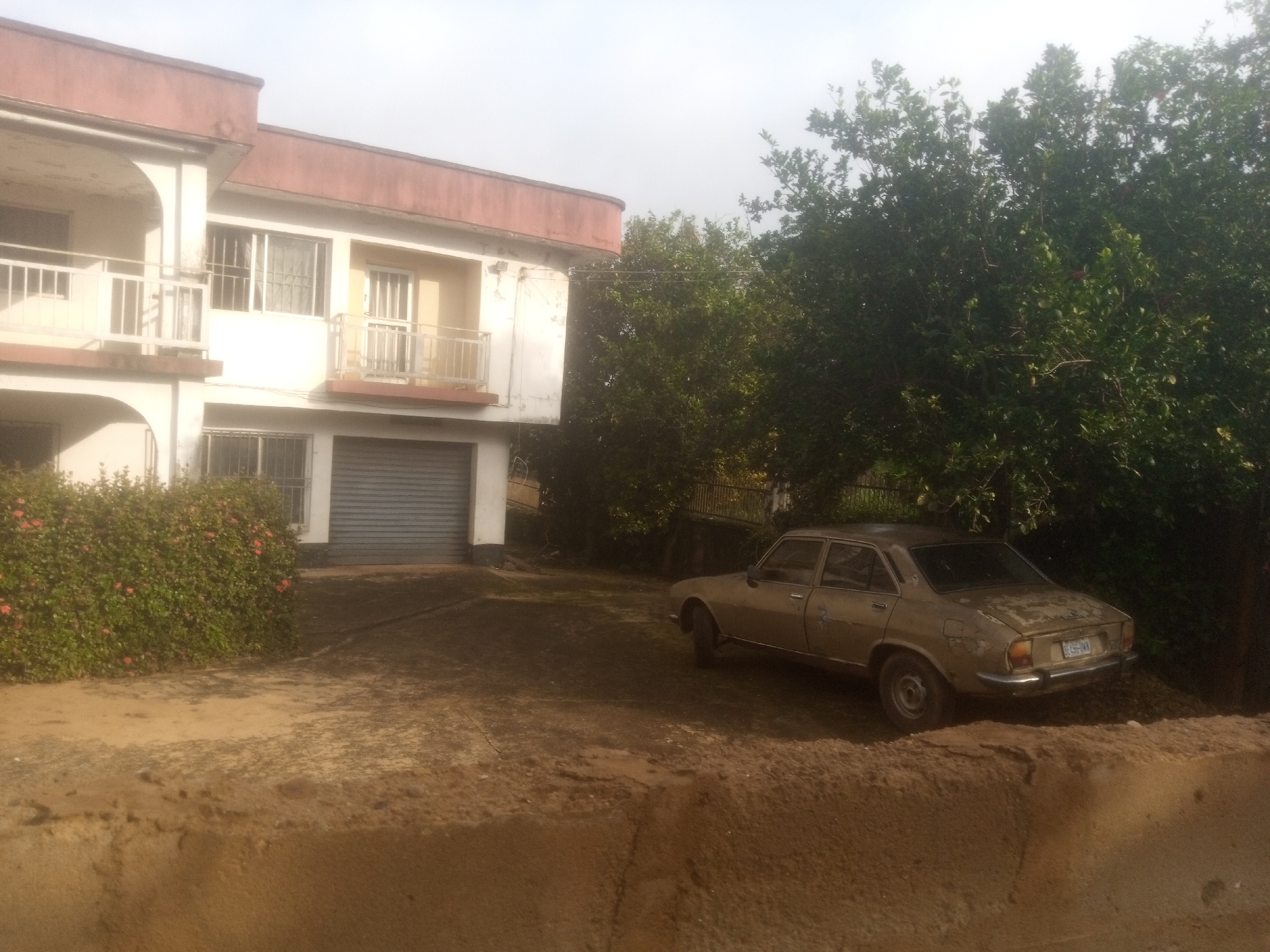 In Nanka, Nigeria, a light-colored, older model sedan is parked on a dirt surface in front of a two-story building. The building has a reddish-brown roofline and off-white walls with multiple windows and balconies. A gray roll-up garage door is visible on the ground floor. To the left of the building, a dense hedge with small orange flowers lines the front. Tall green trees are situated behind the building and to the right, partially obscuring the sky, which appears overcast. The ground is predominantly composed of dirt and patches of concrete.