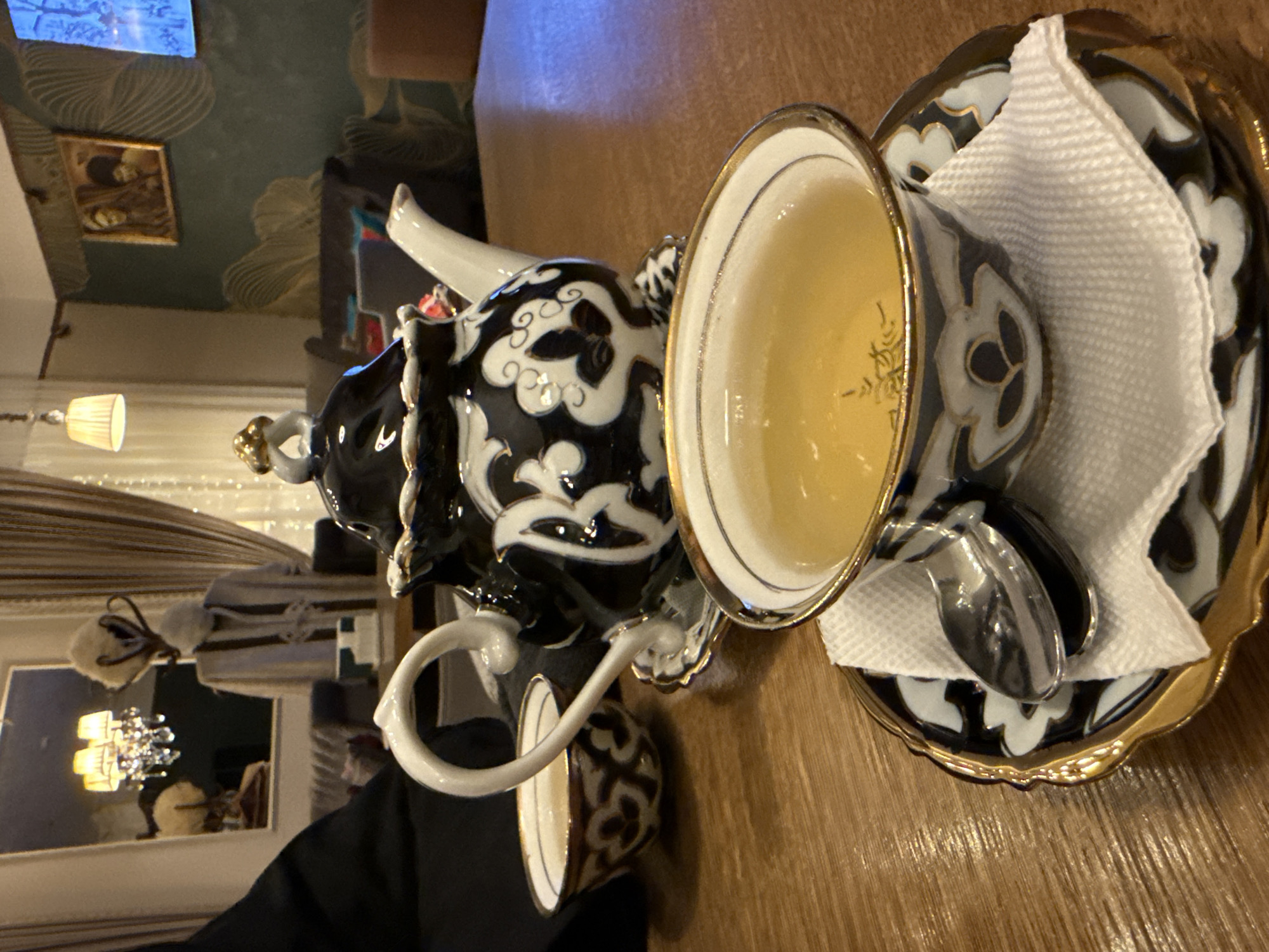 A teapot and teacup set is on a wooden table. The set is black and white with gold trim. The cup is filled with tea. A small spoon rests on a napkin atop the saucer. The table appears to be indoors, with a living room visible in the background featuring a decorative wall and a chandelier. It may be located in Moscow, Russia.