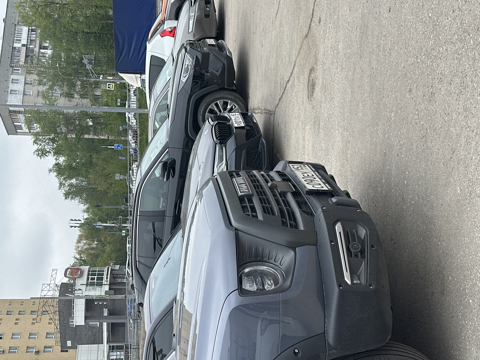 The image depicts a street scene in Nizhny Novgorod, Russia, with a row of parked cars along the right side. The vehicles include a gray Haval SUV with license plate C906C, a black BMW with license plate P001TA, and at least two other vehicles partially visible further down the line. The cars are parked on a paved surface.
In the background, there are buildings, trees, and streetlights. In the left side of the frame, a building with a 'TM' sign on top of it is visible. Traffic lights can also be seen in the background. The sky appears overcast.