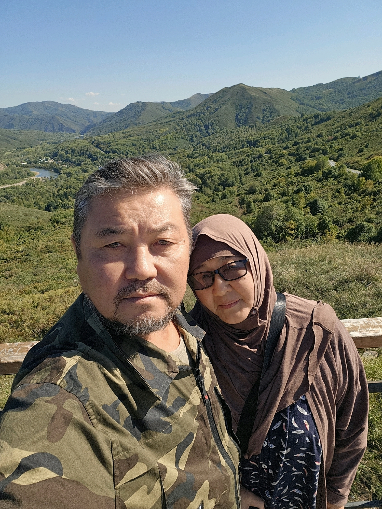 An adult man and woman are taking a selfie outdoors on a sunny day, with a picturesque mountain and forest landscape in the background. The man, in the foreground, has short, graying hair and a goatee. He is wearing a camouflage jacket over a tan shirt. The woman, to his right and slightly behind him, is wearing a brown hijab and glasses, along with a brown open cardigan over a dark blue patterned top. They are standing on a wooden platform or overlook. The background features rolling green hills covered in trees, with a body of water and some structures visible in the valley below. The sky is clear and blue. The location is identified as Tarkhanka rural district, Kazakhstan.