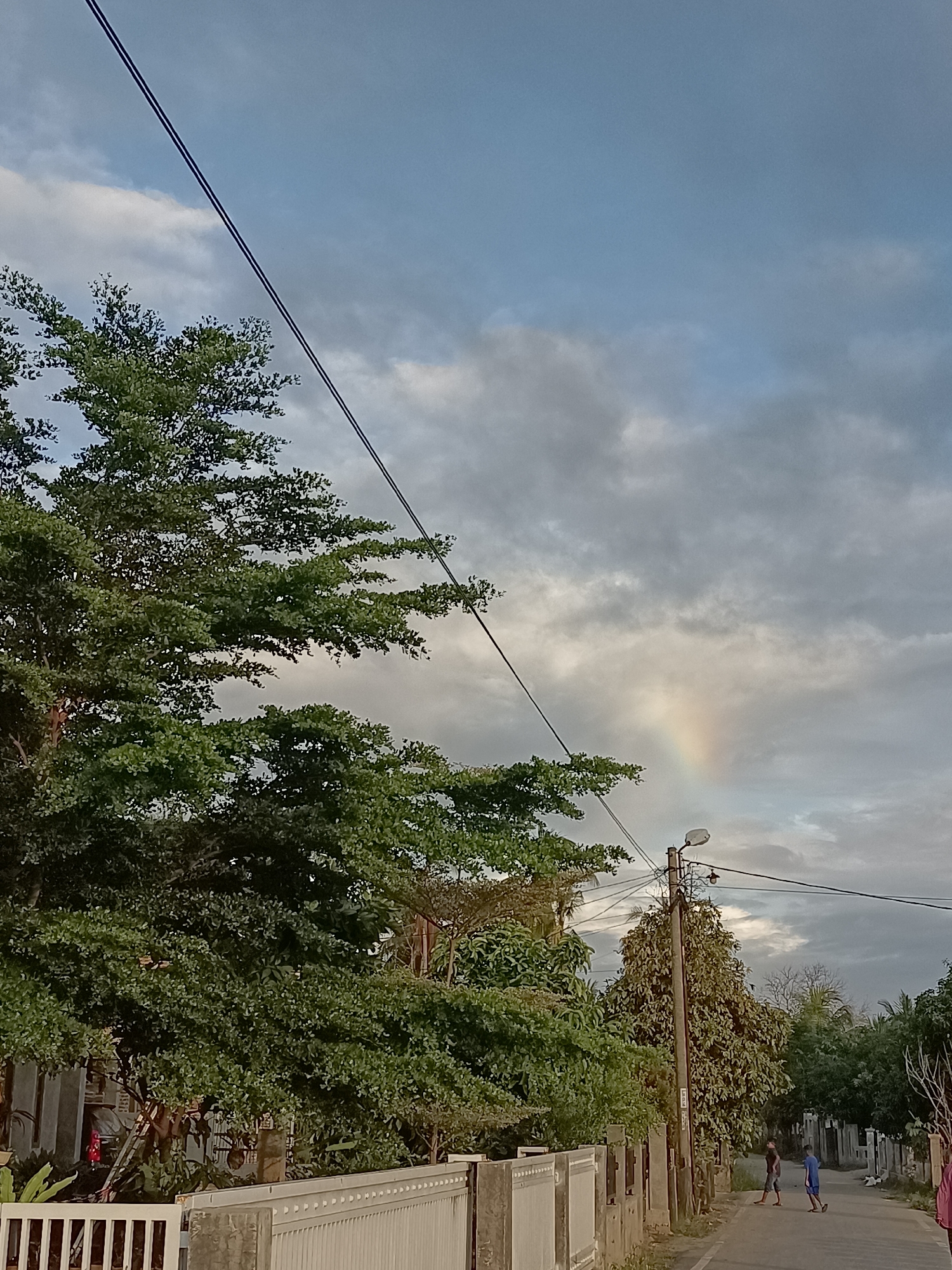 This image depicts a street scene in Meunasah Raya, Indonesia, under a cloudy sky with a faint rainbow visible in the distance. On the left, a large, leafy green tree dominates the foreground, its branches reaching across the frame. A white fence runs along the property line. A power line stretches diagonally across the sky. A utility pole with a street light stands on the right side of the road. Two young boys are walking down the paved street, one in a dark shirt and the other in a blue shirt. In the background, more trees and houses are visible. The lighting suggests it is late afternoon or early evening.