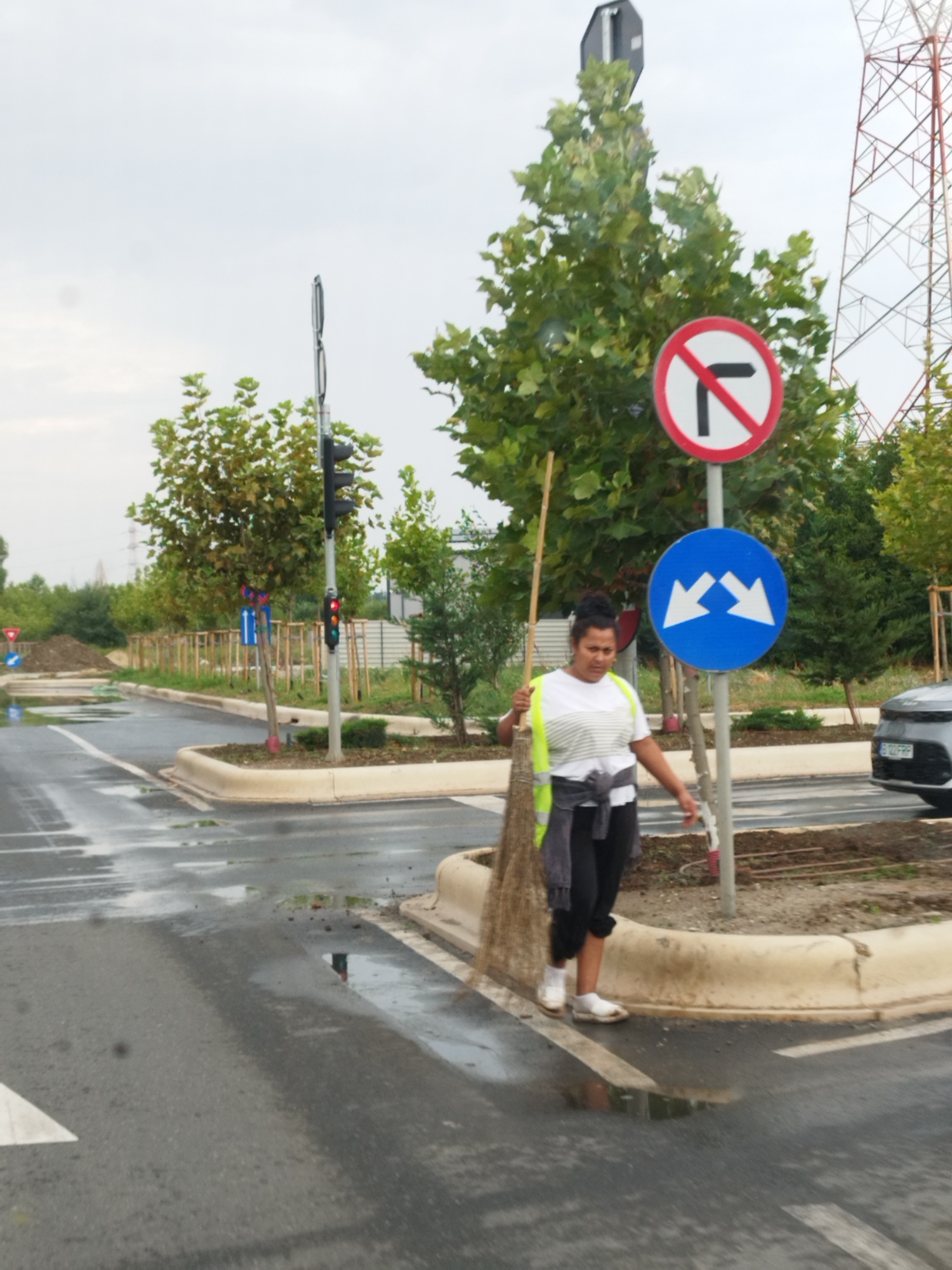 In Bucharest, Romania, a woman sweeps the wet pavement near a street intersection with a broom. She is wearing a white t-shirt with a reflective yellow vest over it, dark pants, and white shoes. The street is wet from rain, with puddles reflecting the overcast sky. Traffic signs are visible: a "no right turn" sign is posted above a blue sign with two white arrows pointing diagonally towards the center. A traffic light with a red light illuminated is also present. A silver car is parked to the right of the scene. Trees with green foliage line the street, and a large metal tower is visible in the background. The ground is a mix of asphalt, dirt, and concrete curbing.