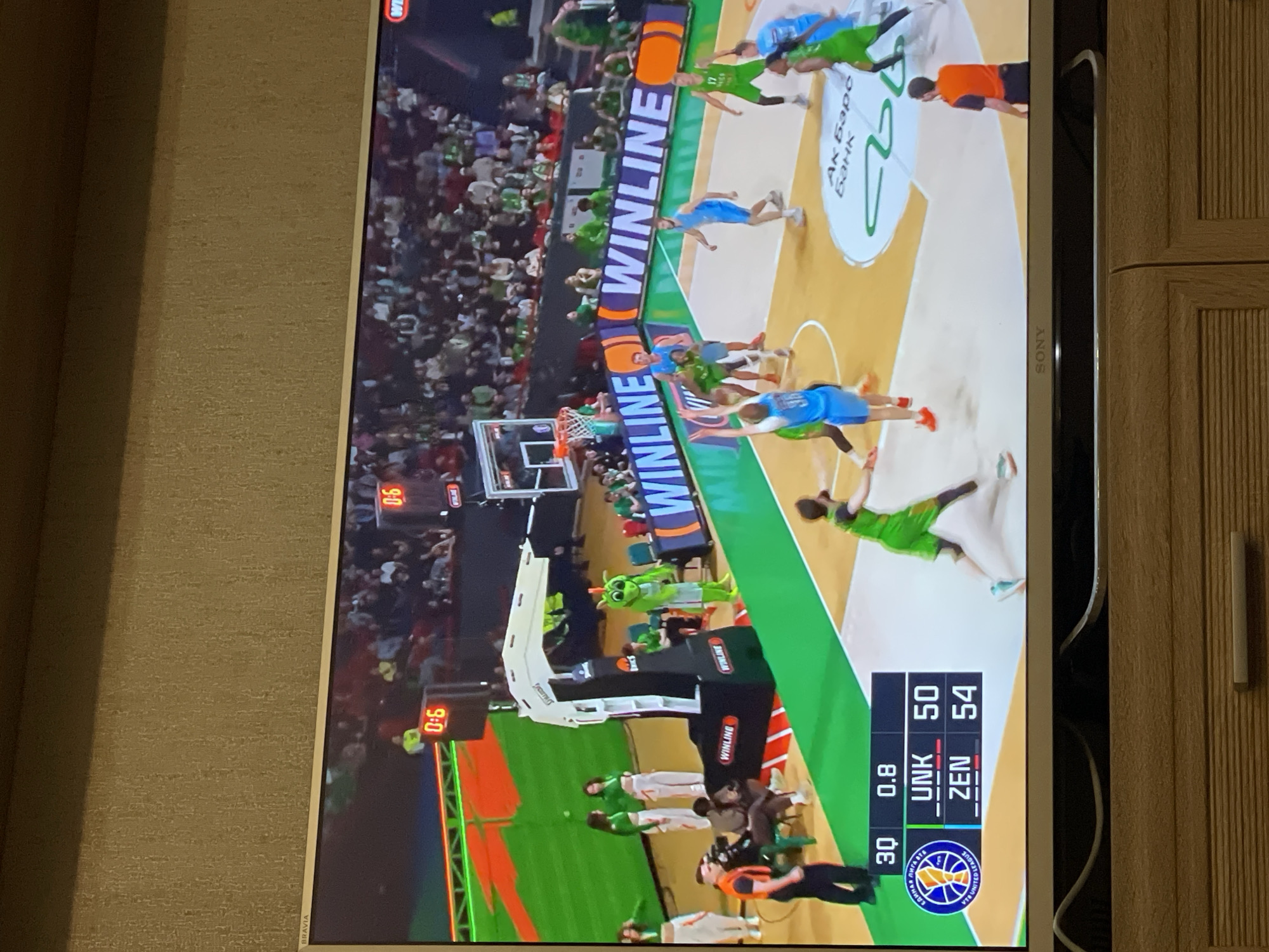 The image shows a basketball game being televised on a Sony television. The game is between UNK (Unics Kazan) and ZEN (Zenit Saint Petersburg) with a score of UNK 50 and ZEN 54, with 0.8 seconds left on the clock. The WINLINE company sponsors the game. There are spectators in the stands and players on the court in green and blue uniforms. This game is likely taking place in the city of Kazan, Russia.