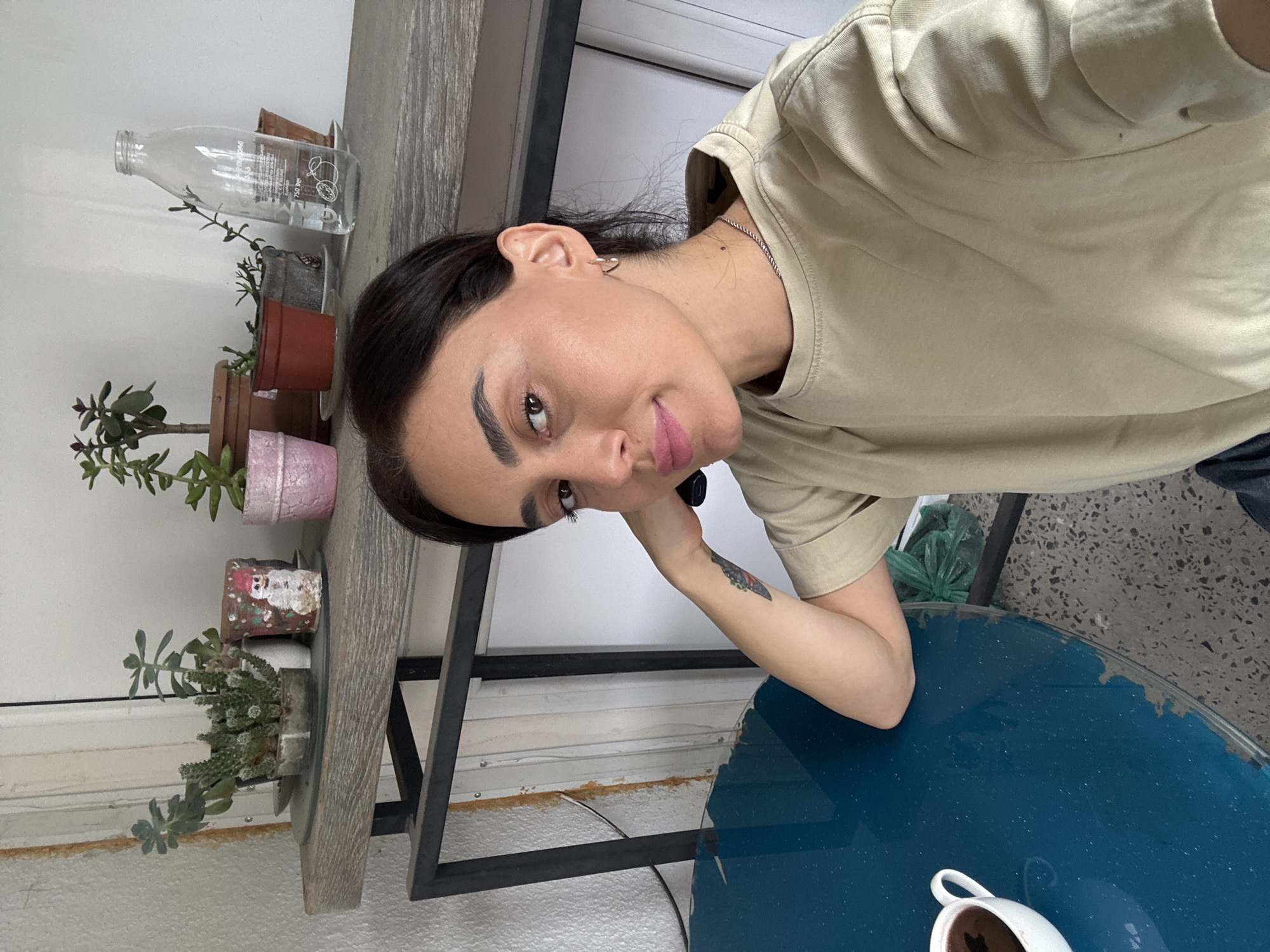 A casually dressed woman with dark hair and light skin is sitting at a blue table with a white cup of dark liquid. She is wearing a light beige t-shirt and has a tattoo on her left arm. The woman is looking at the camera, her head tilted slightly. In the background, there is a shelf with several potted plants and a glass bottle of water. The setting appears to be outdoors, possibly on a balcony or patio, given the rough wall texture and the presence of a metal railing. The overall impression is one of a relaxed, everyday moment captured in a domestic or semi-outdoor urban environment, potentially in Tbilisi, Georgia.