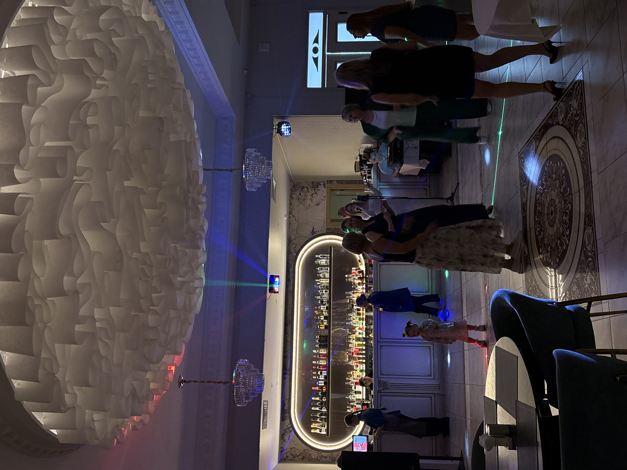 The image depicts an indoor event, likely a celebration or a party, with several people present. In the background, a bar is visible, stocked with numerous bottles of alcohol. Three chandeliers are suspended from the ceiling, along with a modern, textured ceiling design. Colored lights, including red, green, and blue beams, are projected throughout the space, creating a festive atmosphere.
Several individuals are standing and dancing, with some gathered around the bar. A young girl, with her hair in pigtails, is standing in the foreground. Two tables with checkered tops are visible in the lower left corner, along with chairs. The flooring is tiled with an ornate, circular pattern. The overall ambiance suggests a lively social gathering.
The image does not provide any specific clues or identifiable landmarks to determine the city as Birobidzhan, Russia.