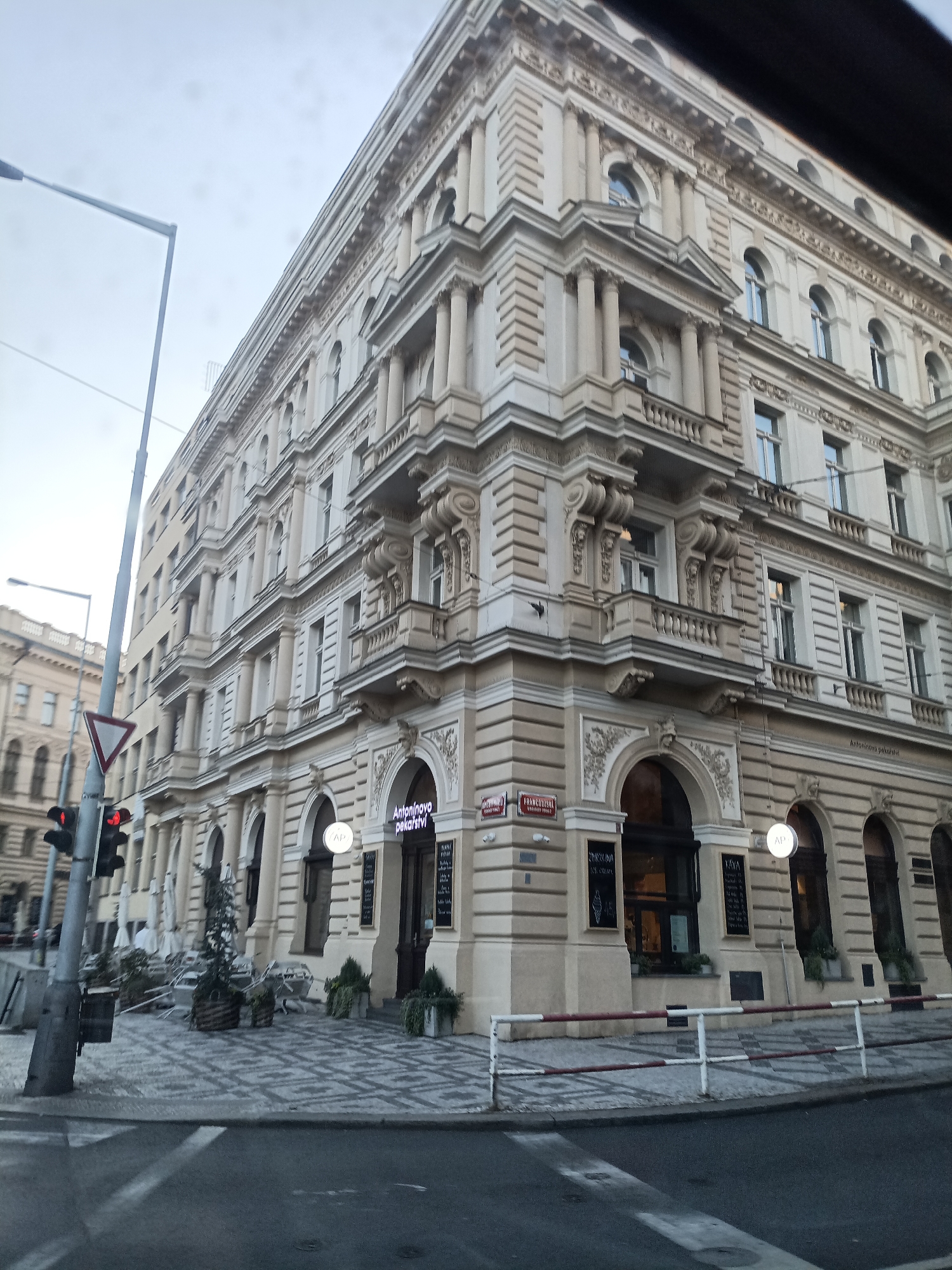 The image shows a street corner in Prague, Czechia, featuring a large, ornate building with a bakery and cafe on the ground floor. The building is beige with elaborate architectural details, including columns, balconies, and arched windows. In front of the building, there are planters with small trees and a few outdoor tables and chairs, suggesting a dining or seating area for the establishment. A traffic light with red and yellow lights is visible on the left side of the image, indicating an intersection. The street is paved with cobblestones and has a pedestrian crossing marked with white lines. The sky is overcast, and the overall lighting suggests it is daytime, possibly late afternoon or early morning due to the muted light. There are no people clearly visible in the frame. The bakery has signage indicating "Antonínovo pekařství" which translates to "Antonín's Bakery."