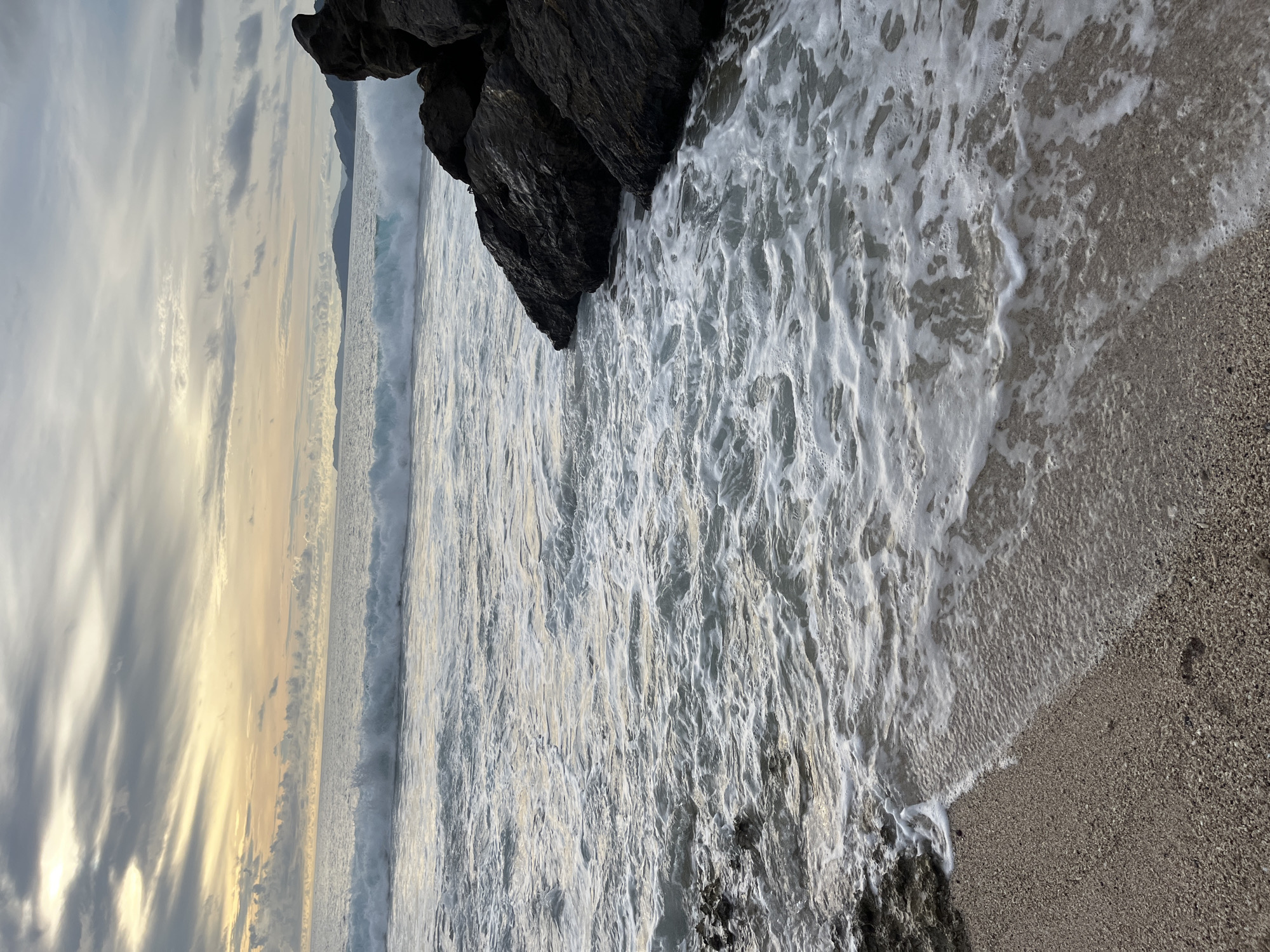 The image captures a dramatic ocean scene with a rough sea crashing against dark, jagged rocks. The perspective is from a slightly elevated viewpoint looking down and across the water.
The main subjects are the powerful waves, characterized by their white foam and turbulent texture, as they surge towards the shore. Large, dark rocks dominate the upper left portion of the frame, providing a stark contrast to the churning water. The sea itself is a dominant element, appearing restless and full of motion. The shoreline is a mix of dark rocks at the bottom and a coarse, pebble-like sand to the right, where the foamy waves are beginning to recede.
The sky above is a significant element, filled with a dramatic display of clouds. Streaks of golden light, suggesting either sunrise or sunset, pierce through the overcast sky, casting a warm, ethereal glow over the horizon. This interplay of light and shadow creates a sense of atmospheric depth and perhaps a touch of melancholy or awe.
The activity is the relentless motion of the sea; waves are actively breaking and washing ashore. The overall mood is one of natural power and wild beauty, with the dynamic interplay of rock, water, and sky evoking a sense of the untamed. The time of day is likely dawn or dusk due to the quality of the light. No people or specific objects are visible, emphasizing the raw, natural landscape. There is no visible text in the image.