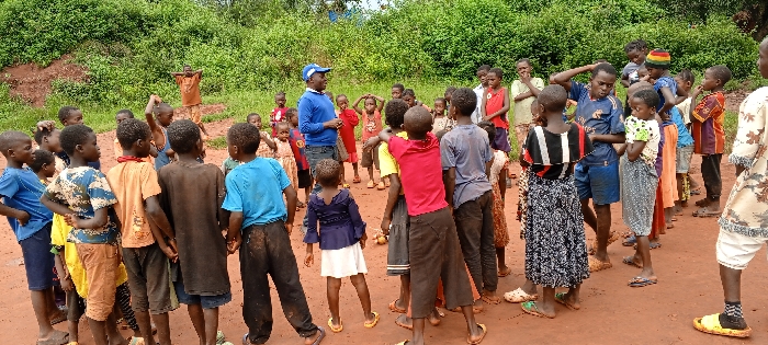 An outdoor scene in Ngalula, Democratic Republic of the Congo, depicts approximately 30 individuals, predominantly children, gathered on a reddish-brown dirt ground. An adult male, wearing a blue long-sleeved shirt and a blue baseball cap, stands in the left-midground, facing a group of children arranged in a loose semicircle. He appears to be addressing or instructing the children. Several children have their hands on their heads or shoulders, suggesting participation in an activity. The children's ages vary, and they wear diverse casual clothing, including t-shirts, shorts, dresses, and skirts of various colors and patterns. Some are barefoot, while others wear sandals or flip-flops. The background consists of dense green vegetation. Small, light-colored, round objects are visible on the ground near the center of the gathering. An additional individual is partially visible on the far right of the frame.