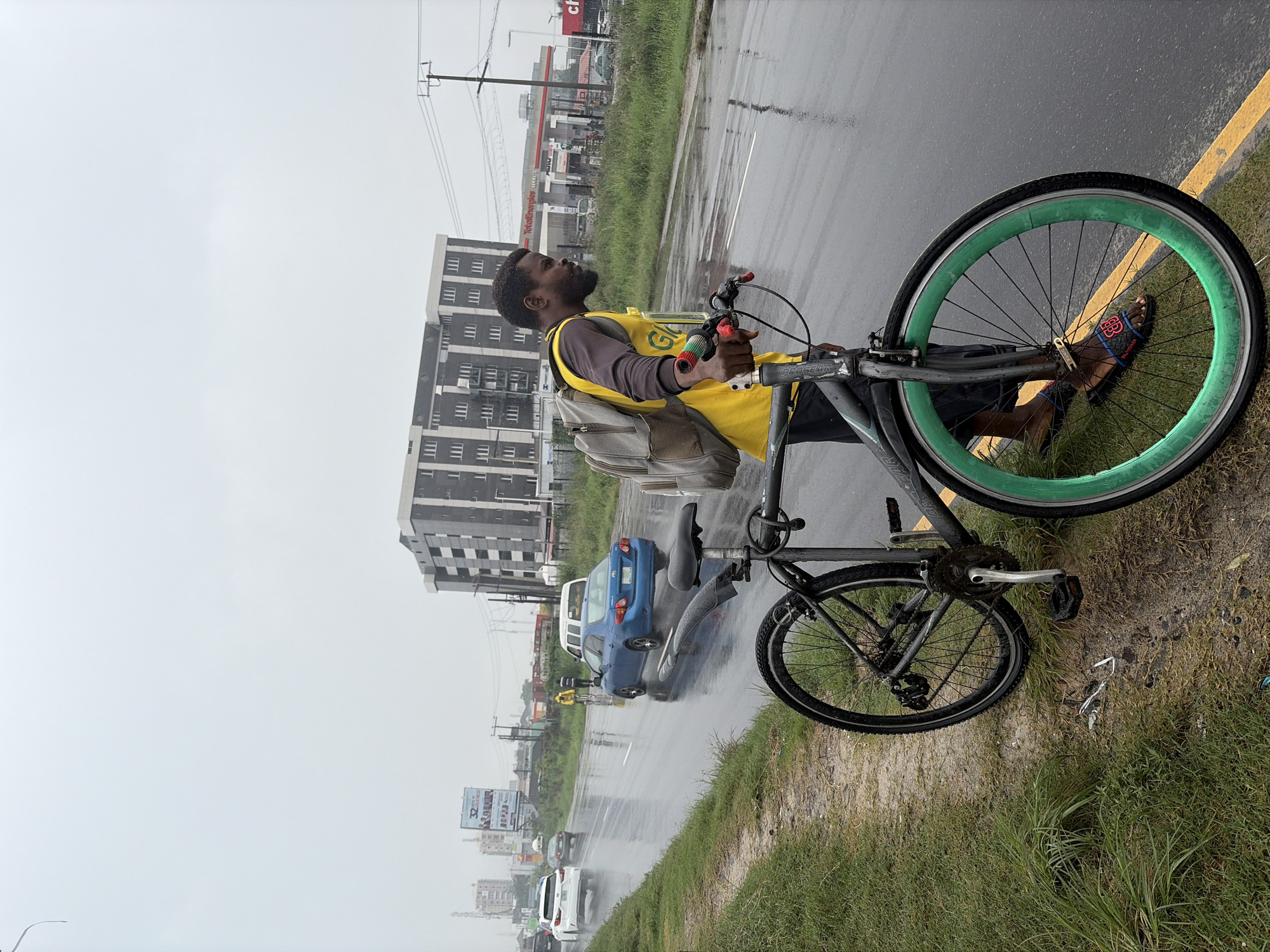 This media file depicts a man standing next to his bicycle on a wet road in Igboefon, Nigeria. The man is wearing a yellow vest over a brown long-sleeved shirt and a grey backpack. His right foot is on the pedal of the bicycle, which has a distinctive bright green rim on the front wheel. He is looking up and to his left.
The scene appears to be a roadside with a paved road, a grassy embankment, and a line of traffic in the background. Several cars, including a blue sedan and a white SUV, are visible on the wet road, suggesting it might be raining or has recently rained. Beyond the road, there are buildings, including a multi-story structure, and what looks like a gas station with the "TotalEnergies" logo visible. Overhead power lines are also present. The overall atmosphere is somewhat overcast, consistent with rainy weather.
Notable details include the man's attire and the vibrant green rim of the bicycle wheel. The presence of vehicles on the wet road indicates ongoing traffic. The background scenery with the buildings and gas station provides location cues. The text visible includes "TotalEnergies" on the gas station and "PROGEAR" on the bicycle frame. The man's posture and gaze suggest he might be waiting or contemplating something. The scene is captured during daylight hours, although the cloudy weather might make it appear dimmer.