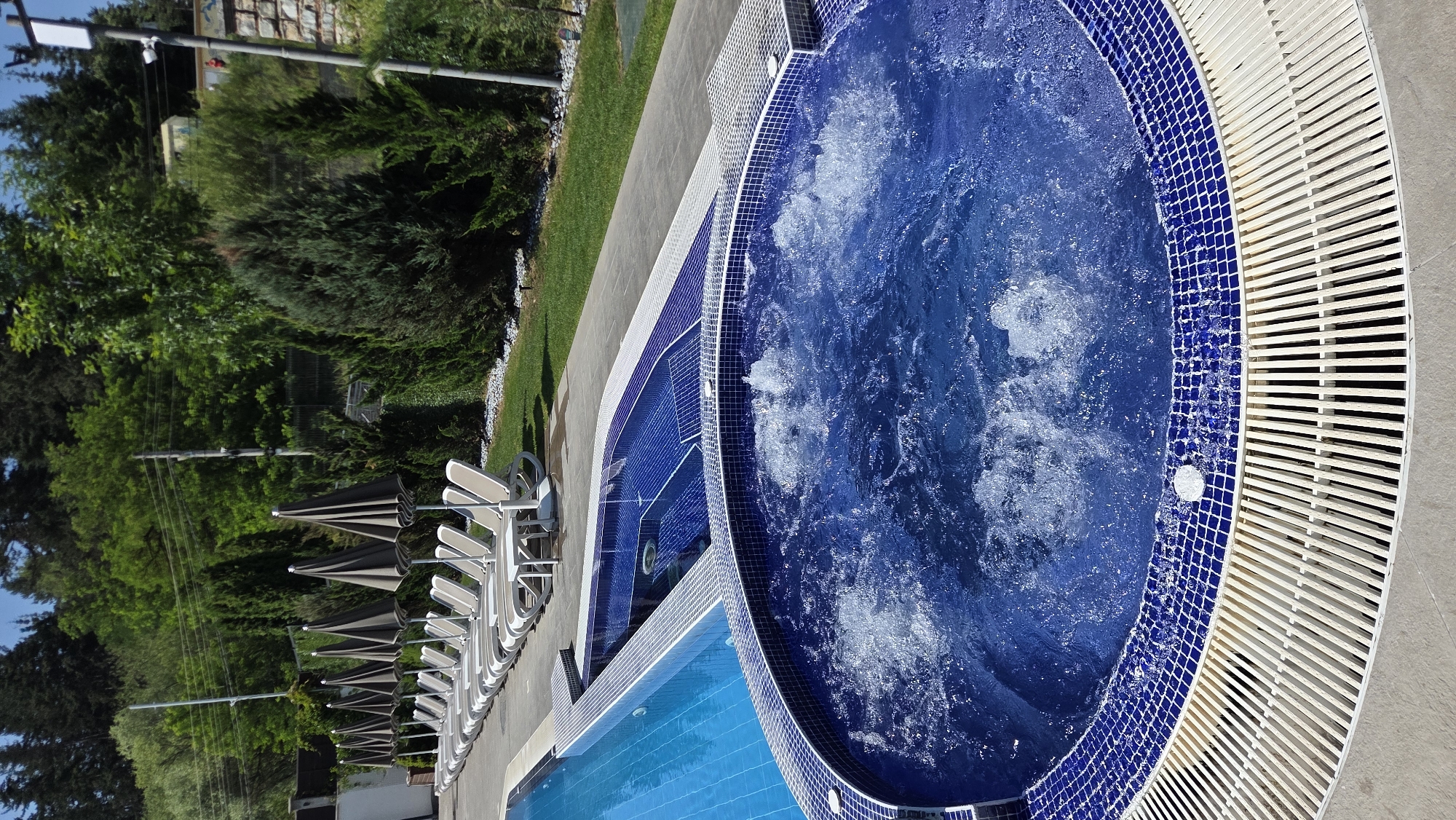 The image shows an outdoor pool area with a separate Jacuzzi in the foreground. The Jacuzzi is circular, filled with bubbling blue water, and has a blue tiled border. Adjacent to it is a rectangular pool, also with blue tiles and a blue tiled surround. Beyond the pools, there is a row of closed lounge chairs and umbrellas, a grassy area, and lush green trees. In the background, there are more trees and some buildings. The scene appears to be set on a sunny day.